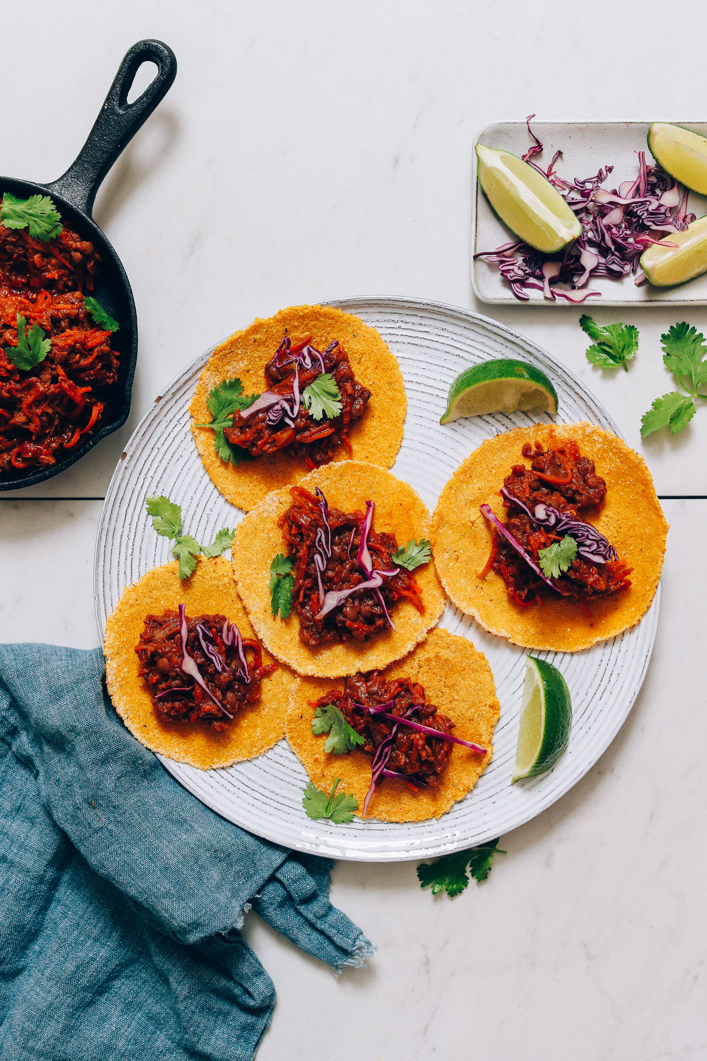Plate of tacos, cilantro, and limes next to a cast iron skillet of vegan lentil taco meat