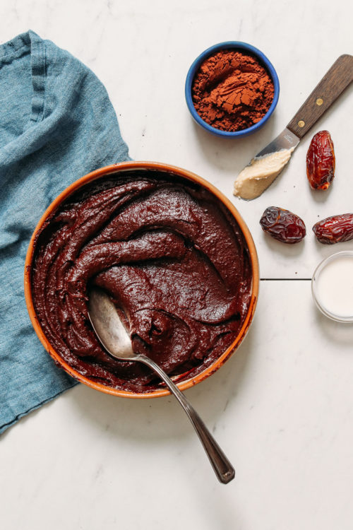 Vintage spoon in a bowl of Vegan Chocolate Frosting beside almond milk dates, cashew butter, and cacao powder