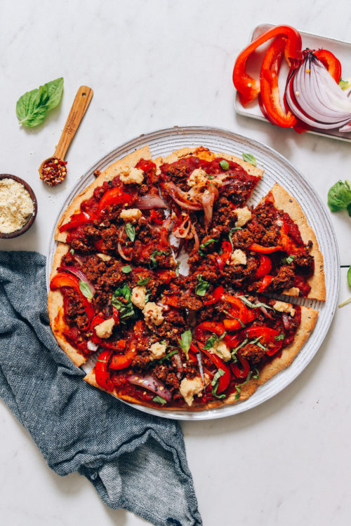 Plate of Vegan Pizza surrounded by red onion, basil, bell peppers, red pepper flake, and vegan parmesan