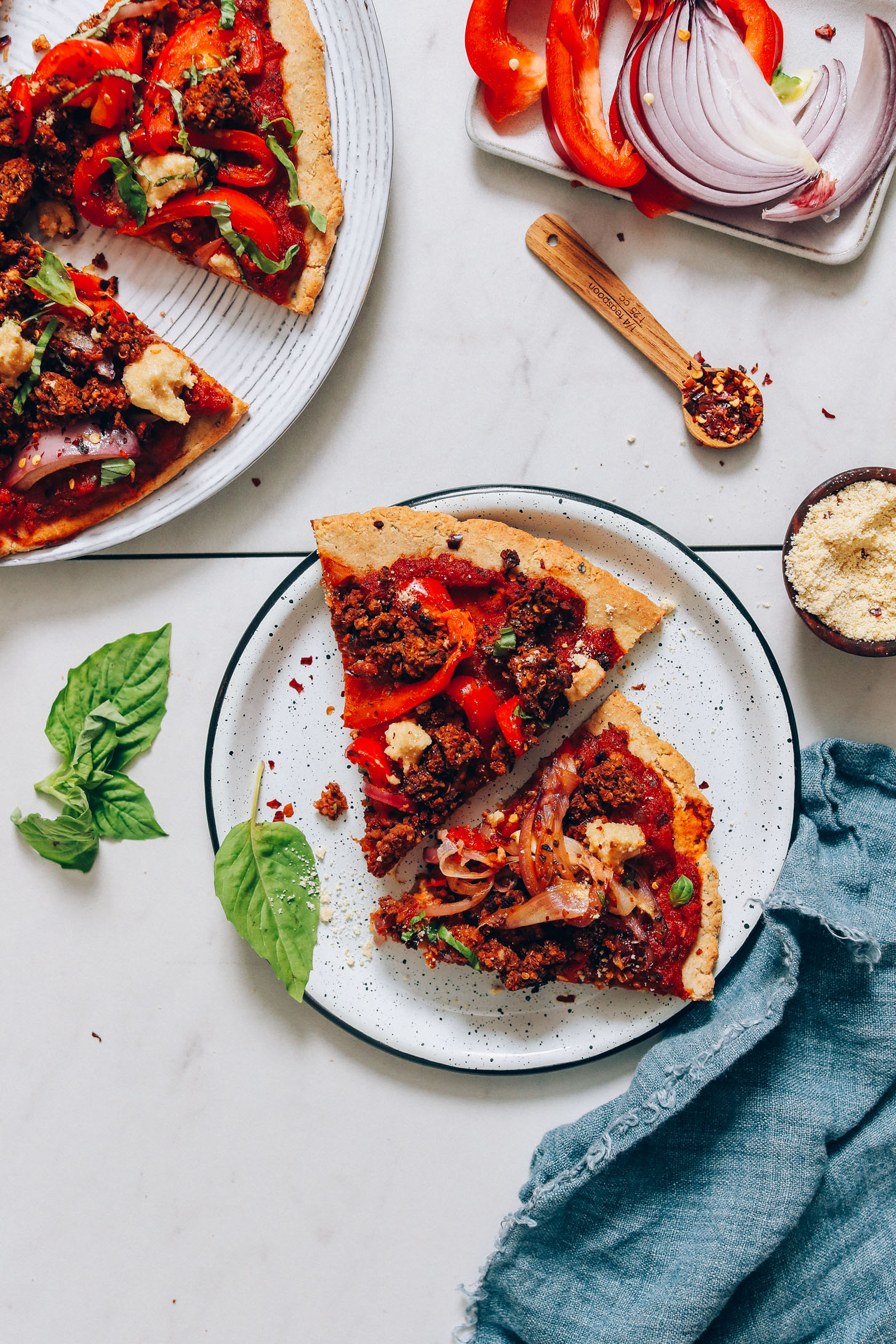Red onion, bell pepper, vegan parmesan, and red pepper flakes next to plates with slices of vegan pizza
