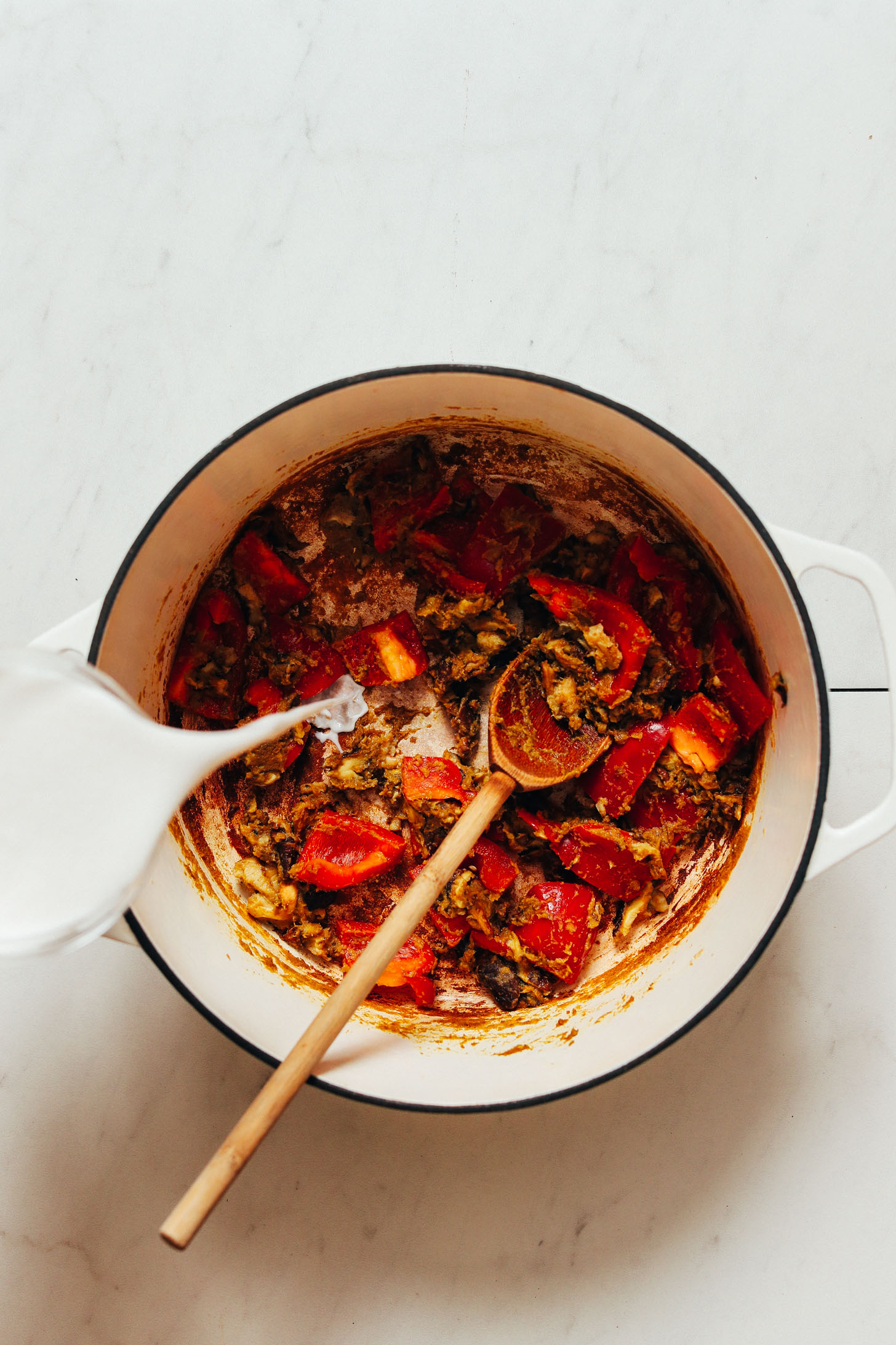 Pouring coconut milk into a pot of bell peppers, mushrooms, and green curry paste