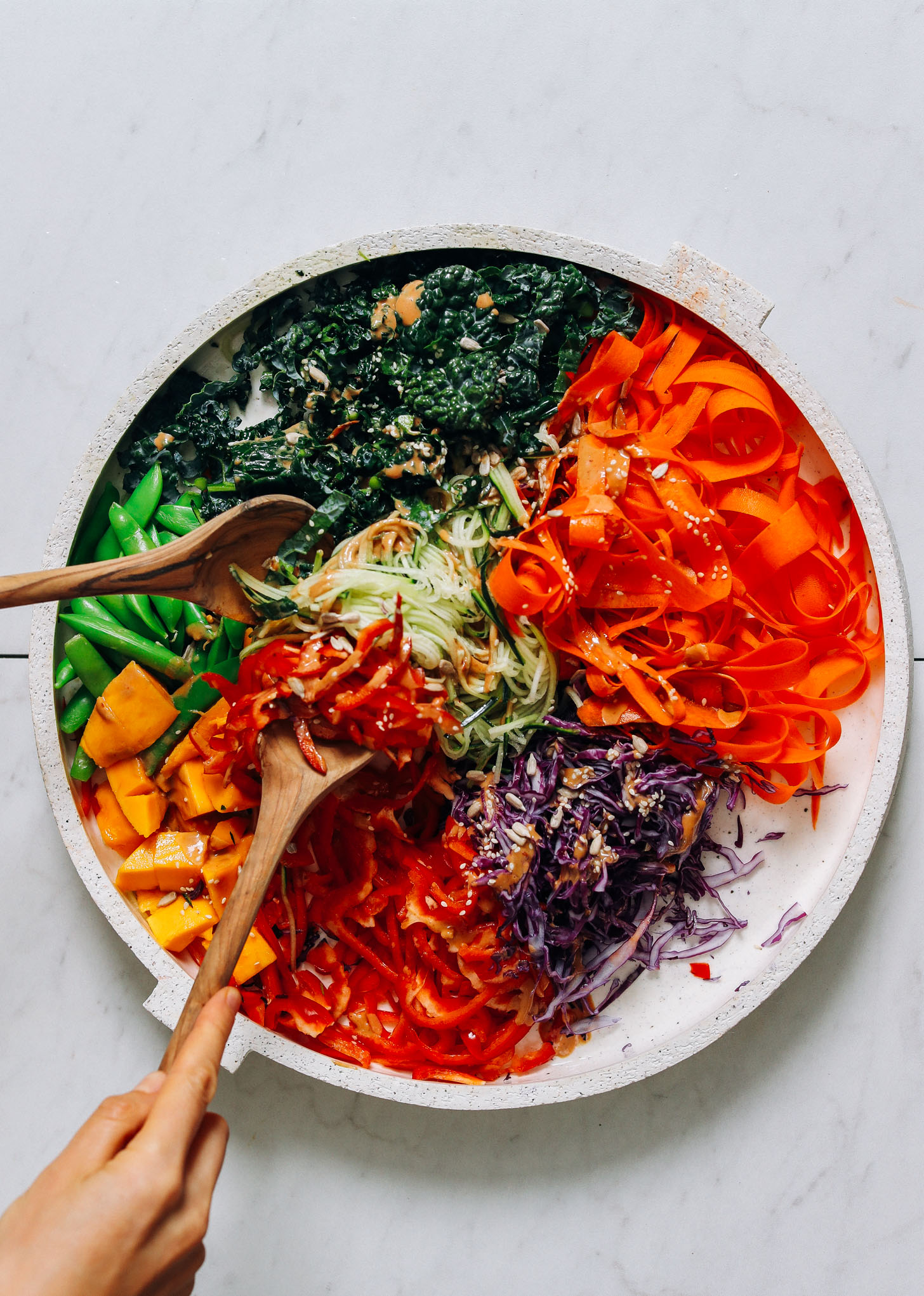 Using salad tongs to mix cucumber noodles, bell pepper, and other vegetables with peanut sauce