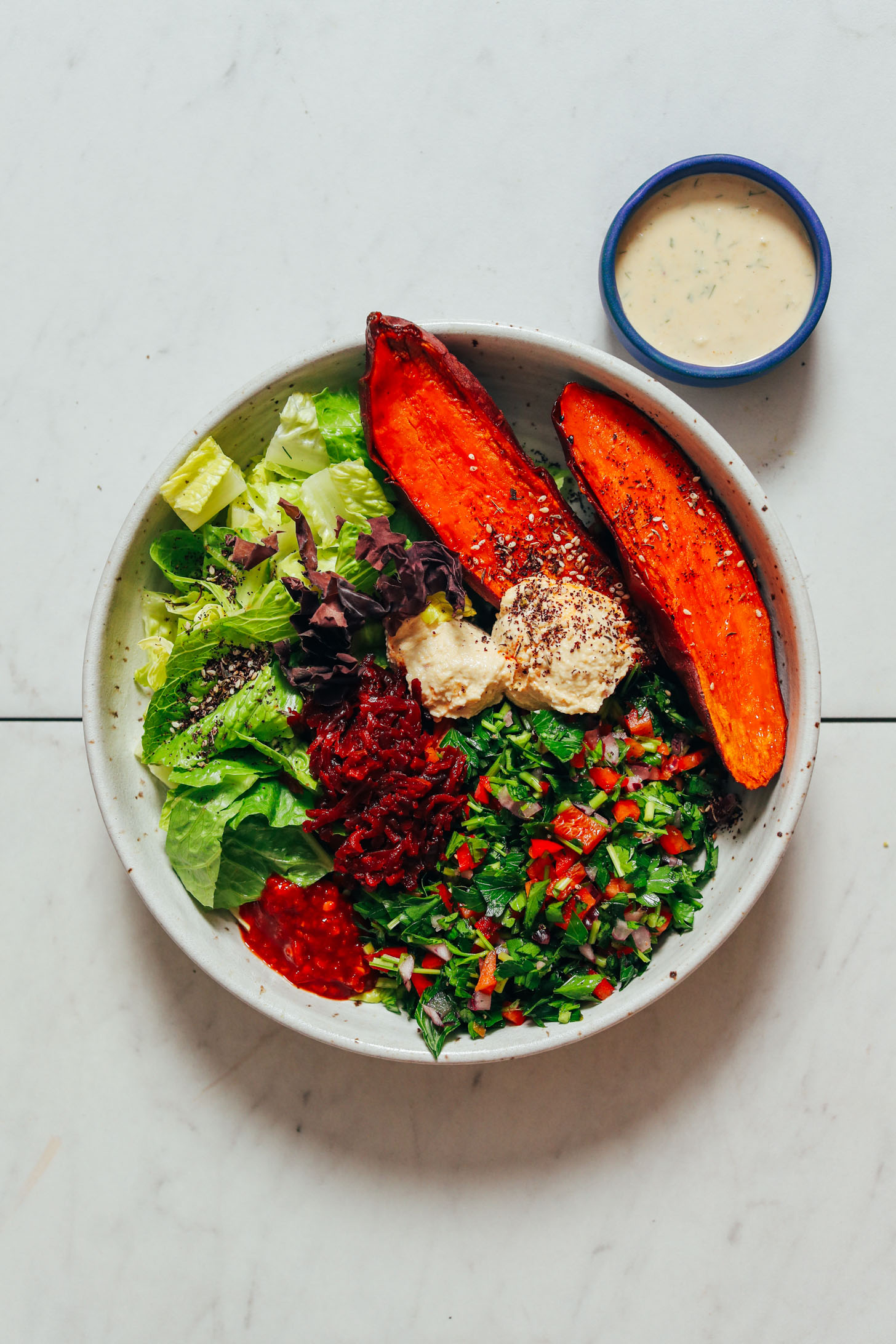 Bowl of dill garlic sauce next to a bowl of sweet potato, hummus, tahini, sauerkraut, parsley salad, chili garlic sauce, za'atar, dulse, and romaine