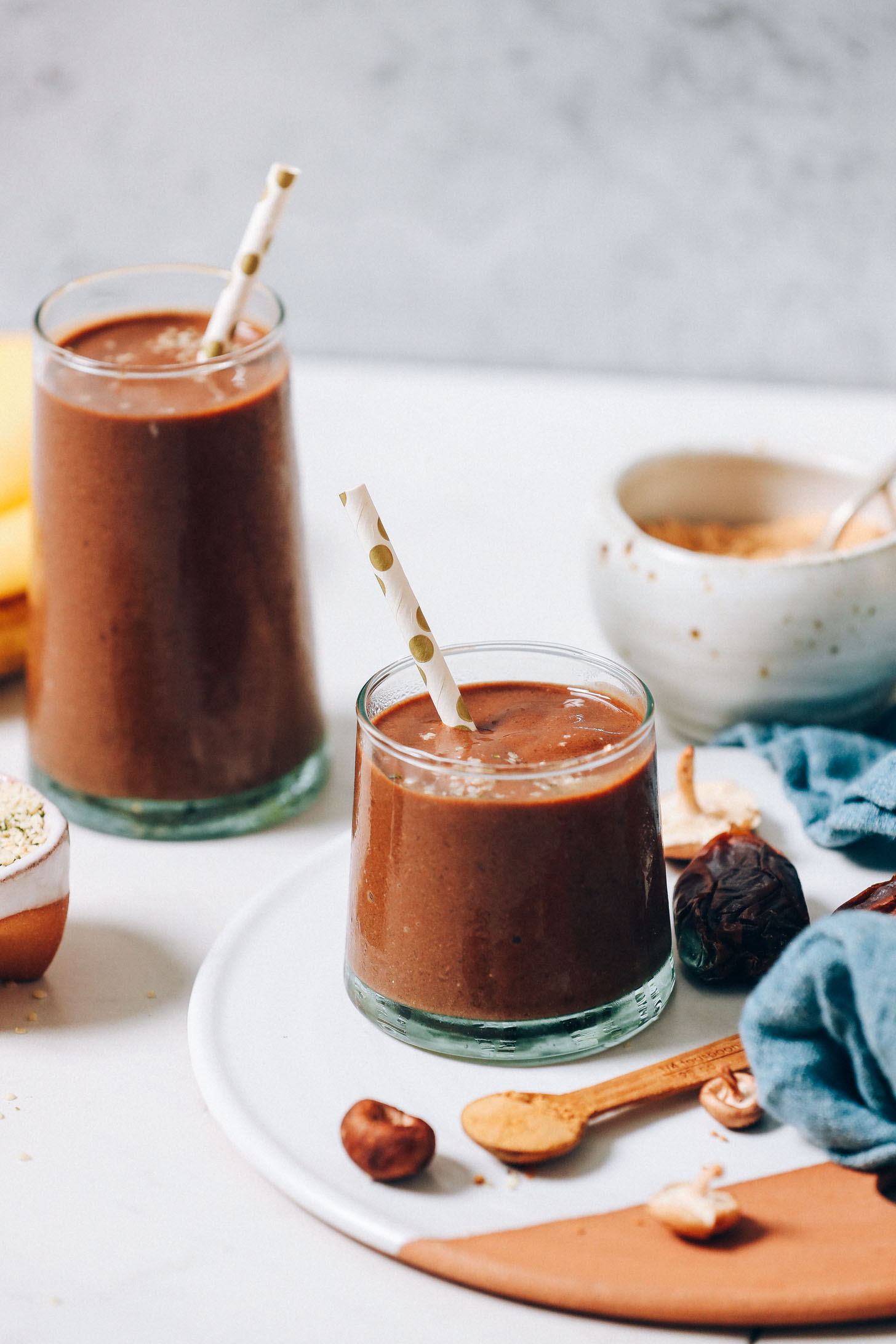 Our adaptogenic chocolate banana shake in large and small glasses next to ingredients used to make it