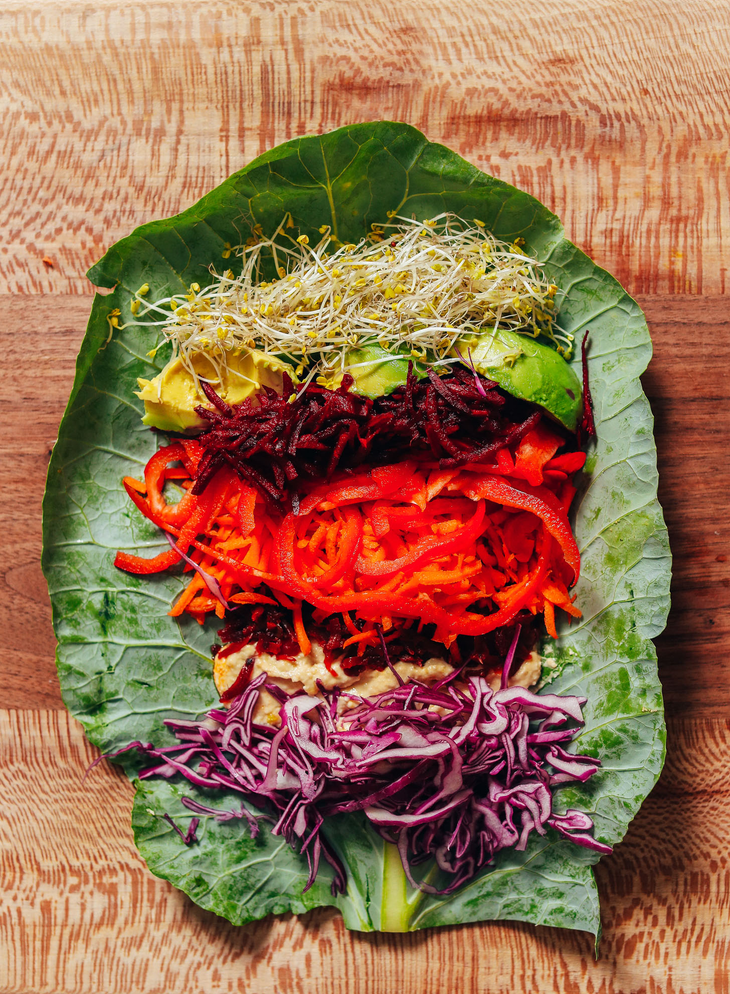 Cutting board with a collard grean leaf topped with broccoli sprouts, avocado, shredded beet, carrot, bell pepper, sauerkraut, hummus, and cabbage