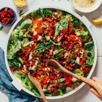 Using wooden tongs to pick up a serving of Vegan Caesar Salad topped with Sweet Potato Croutons