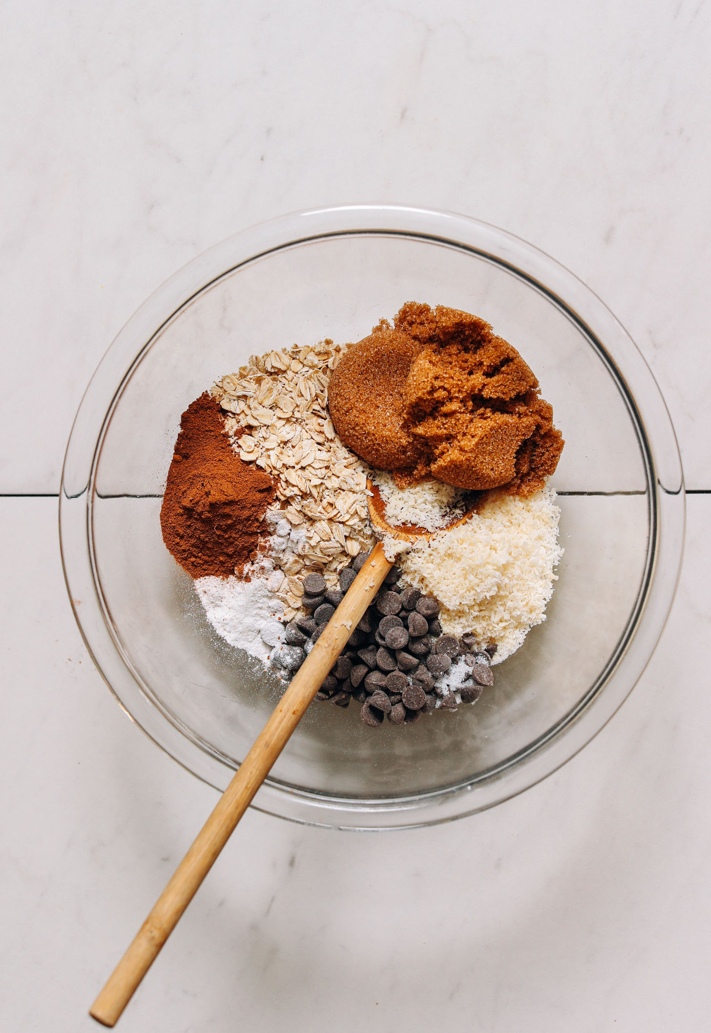 Wooden spoon in a mixing bowl of oats, brown sugar, shredded coconut, chocolate chips, baking powder, and pumpkin pie spice