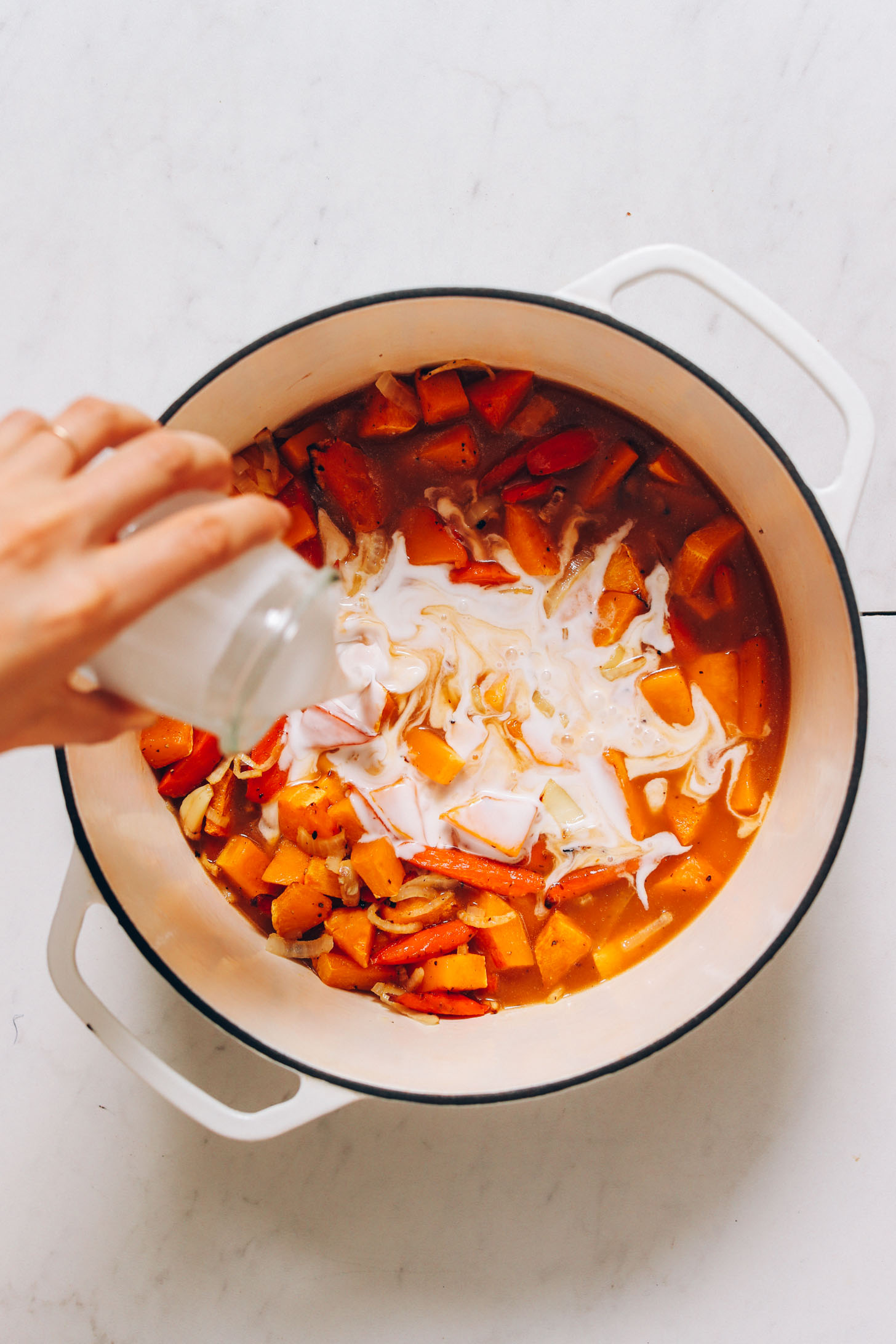 Pouring coconut milk into a pot with carrots, butternut squash, garlic, onion, and vegetable broth