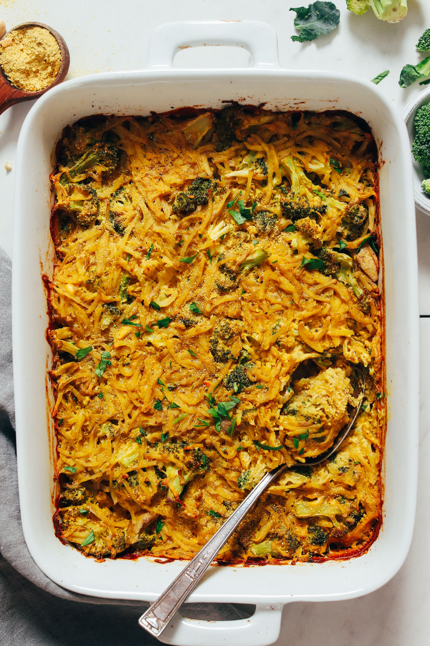 Nutritional yeast and broccoli next to a pan of Cheesy Broccoli Hashbrown Bake