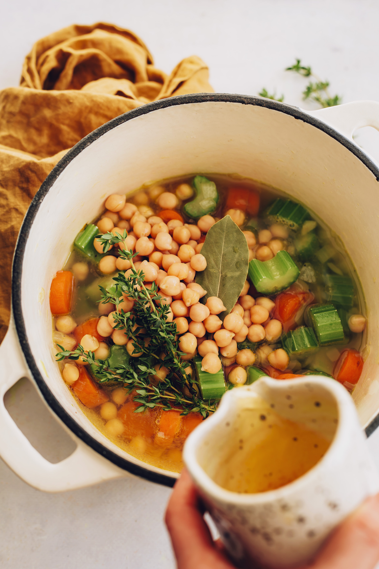 Pouring vegetable broth over carrots, celery, thyme, chickpeas, and a bay leaf