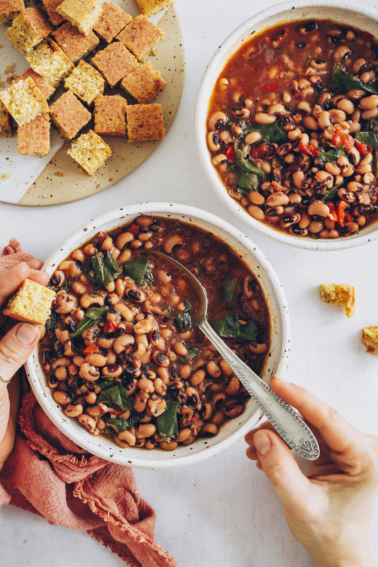 Holding a cube of cornbread above bowls of vegan Black Eyed Peas & Greens