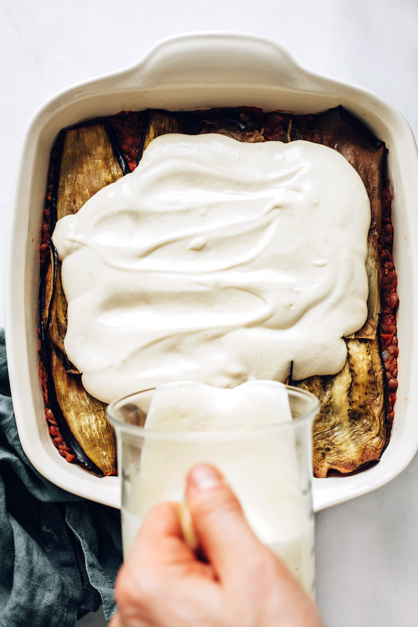 Pouring cashew bechamel sauce over eggplant and lentils to make moussaka