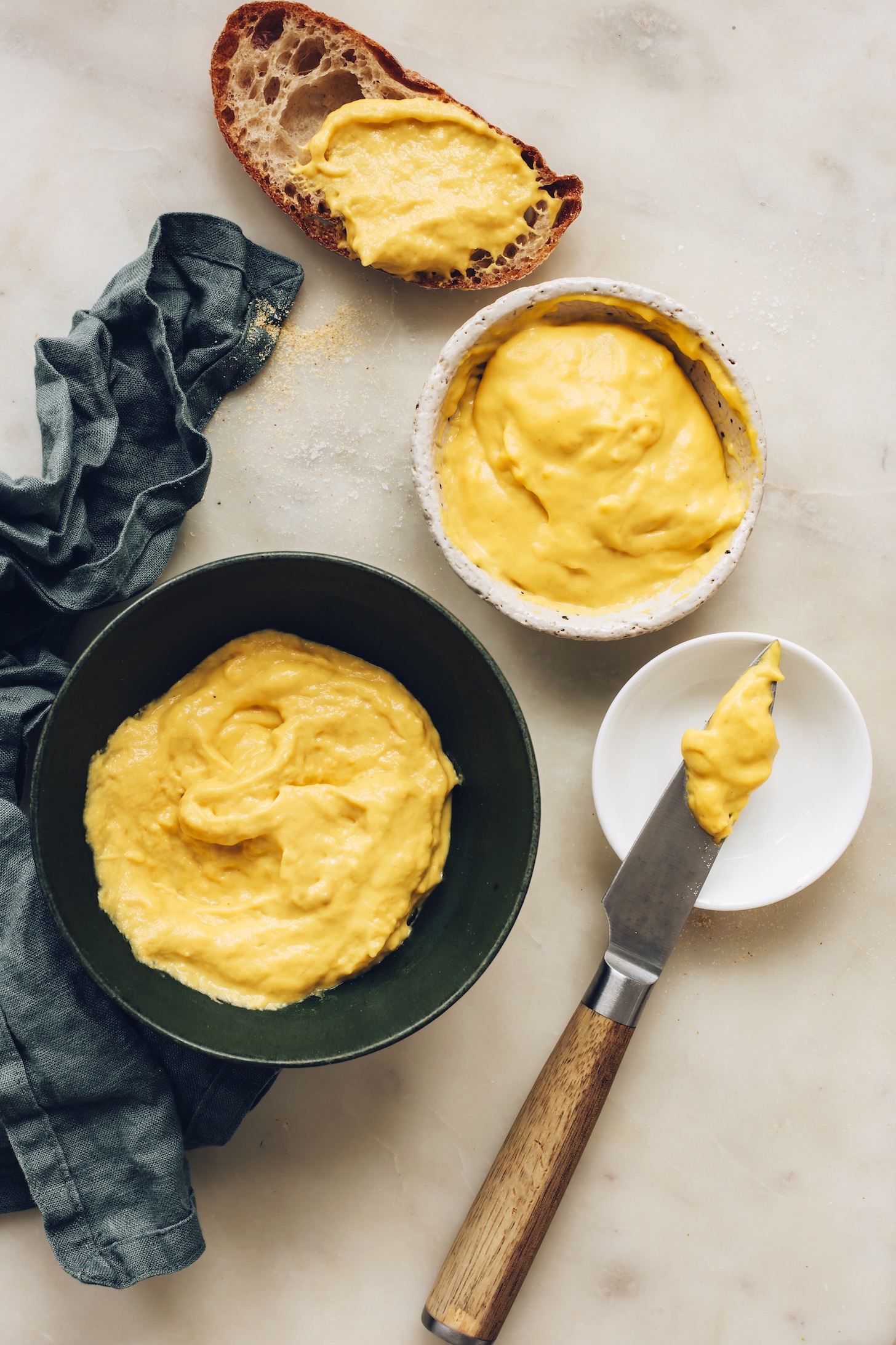 Slice of sourdough bread next to bowls of our easy vegan cheddar cheese recipe