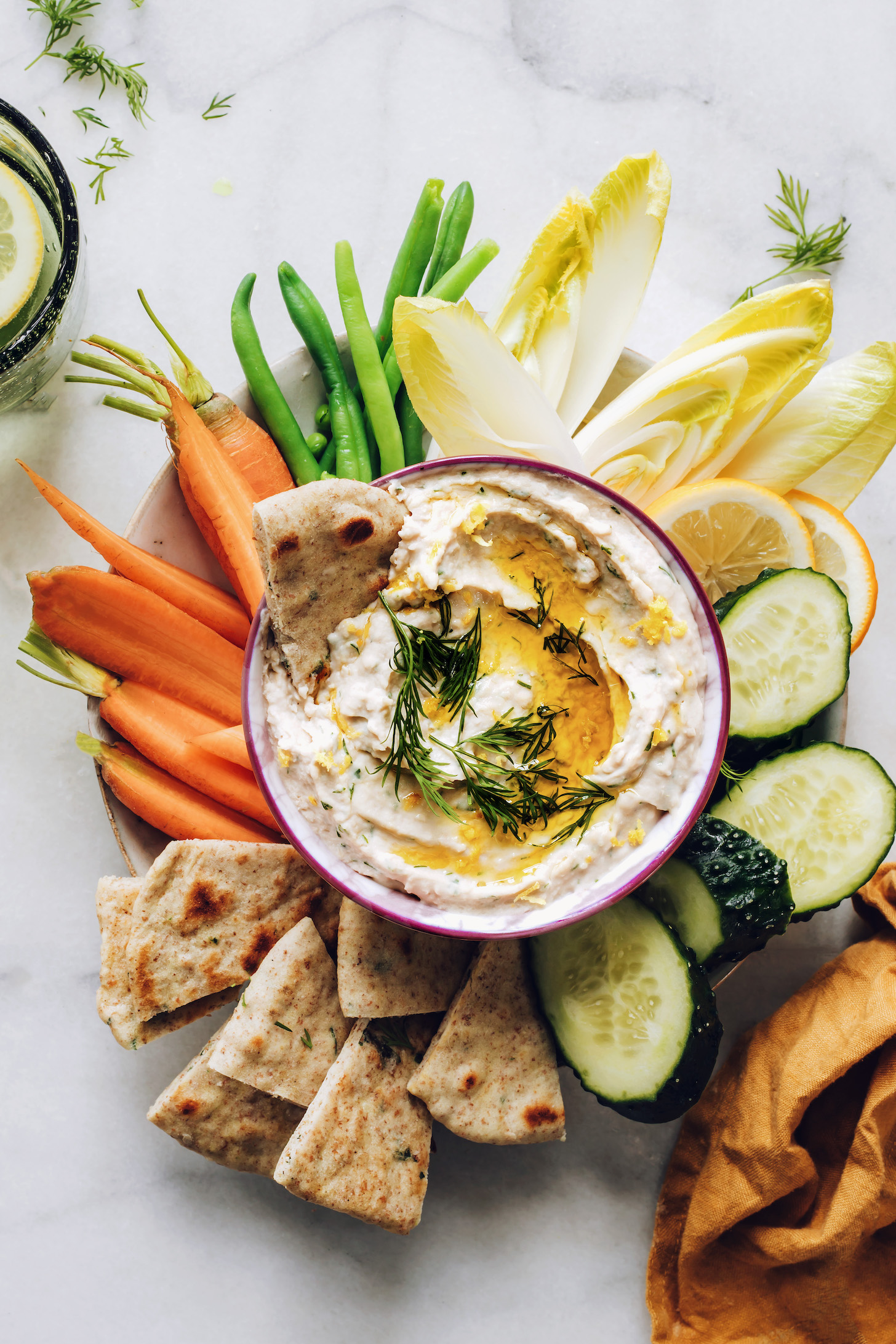 Fresh vegetables and pita bread around a bowl of white bean hummus