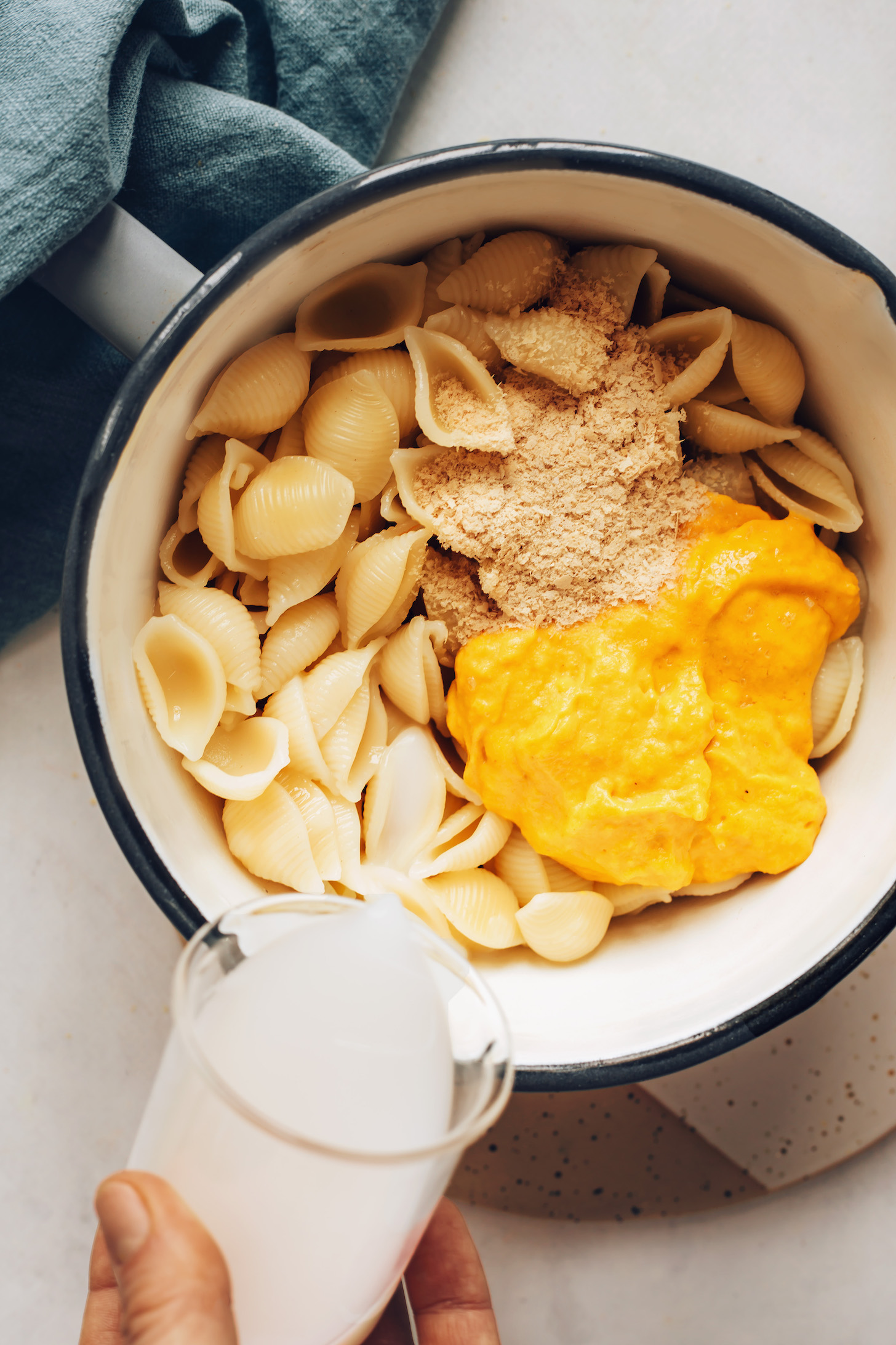Pouring almond milk into a pot of gluten-free pasta, nutritional yeast, and vegan cheddar cheese