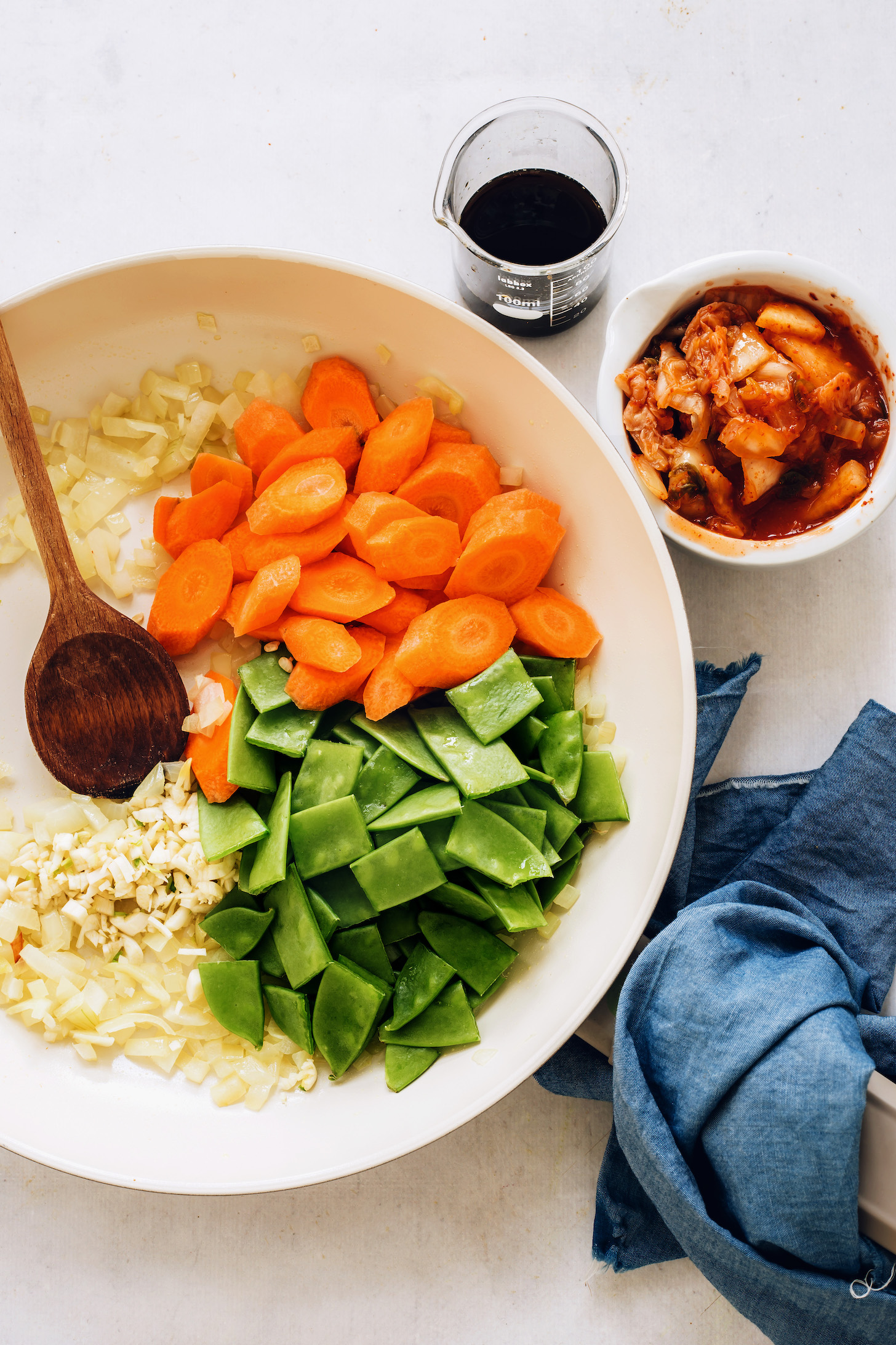 Chopped snap peas, carrots, onions, and garlic in a skillet next to tamari and kimchi