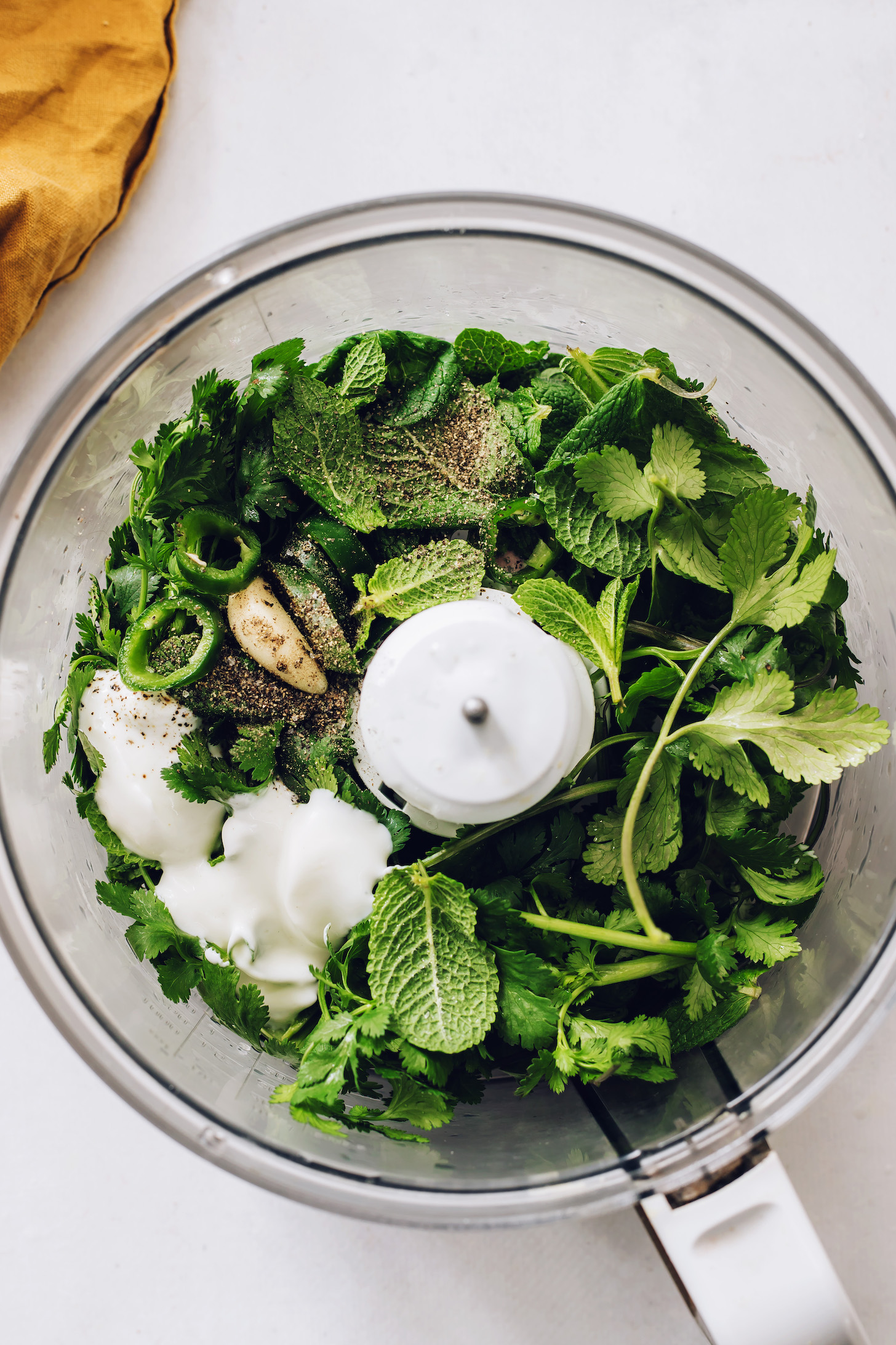 Food processor with mint leaves, cilantro, garlic, jalapeño, lemon juice, maple syrup, coconut yogurt, salt, and pepper
