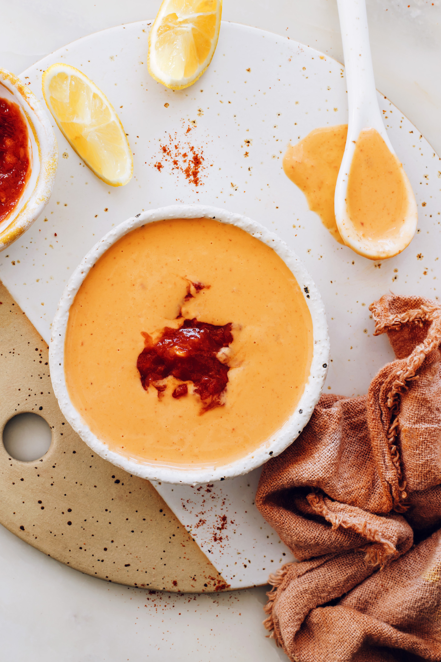 Bowl of harissa tahini sauce next to harissa sauce, a lemon slice, paprika, and tahini