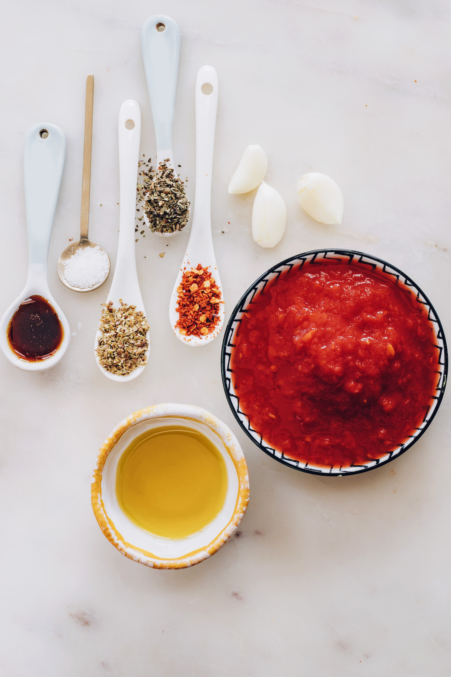 Garlic cloves, spoonfuls of herbs, red pepper flakes, salt, and maple syrup, and bowls of olive oil and crushed tomatoes