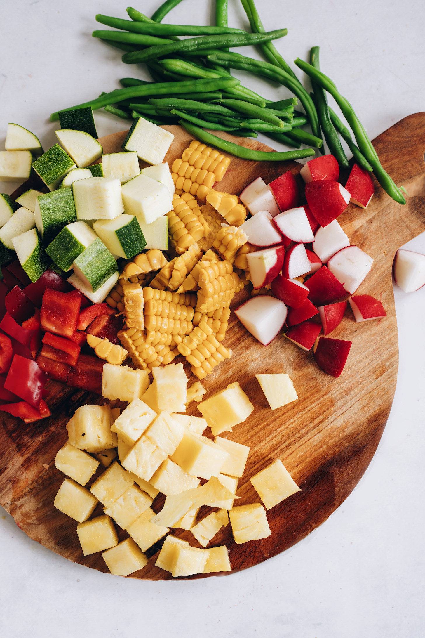 Cutting board with fresh green beans, zucchini, red bell pepper, corn, radishes, and pineapple