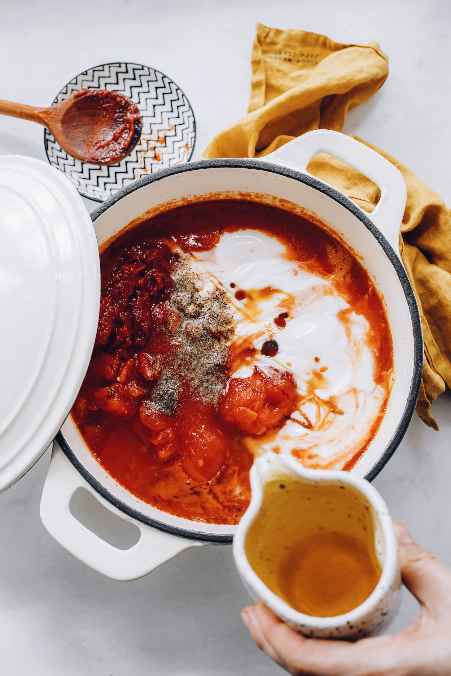 Pouring vegetable broth into a pot with tomatoes, coconut milk, salt, pepper, basil, onion, garlic, maple syrup, and broth.