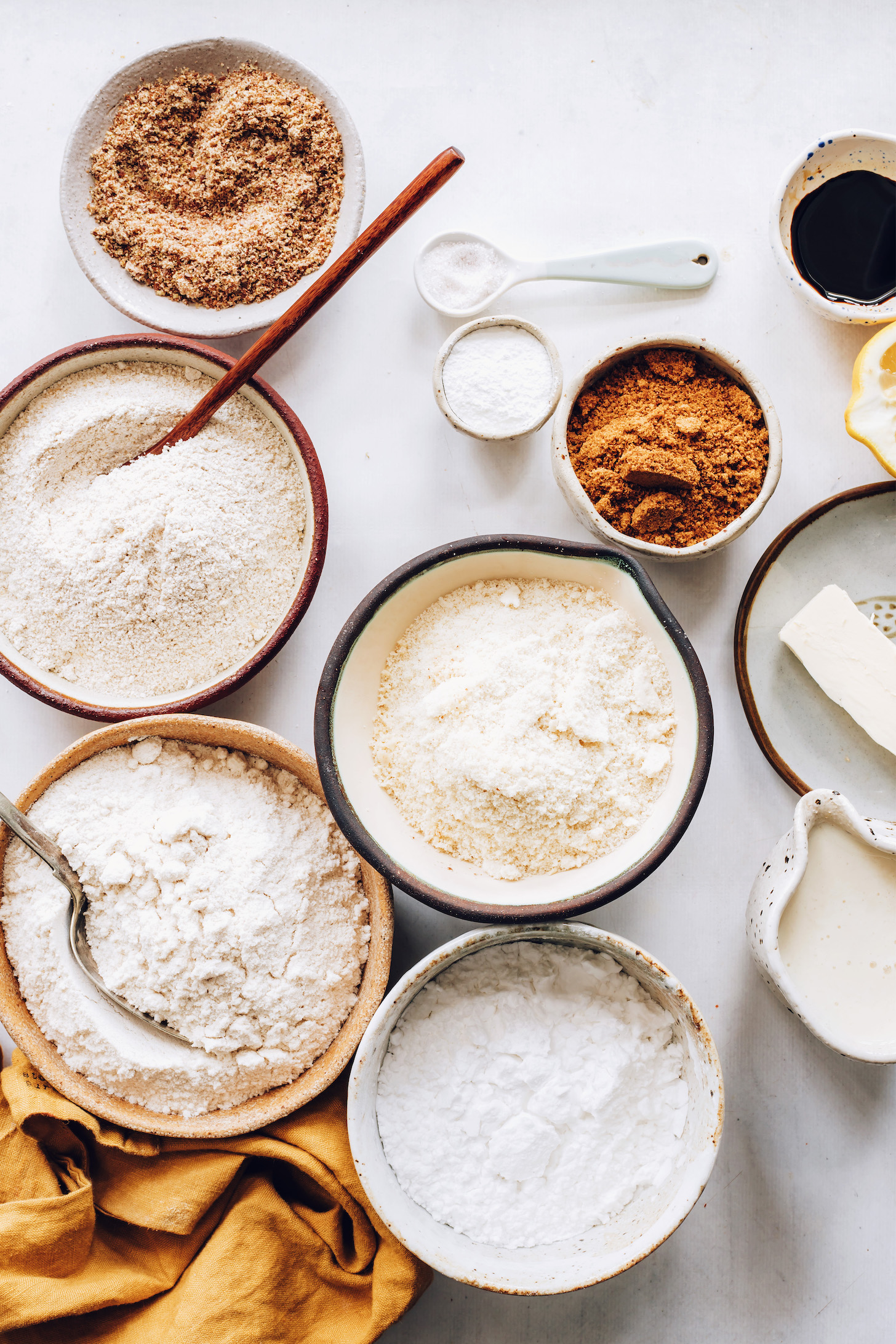 Bowls of brown rice flour, almond flour, oat flour, potato starch, baking powder, flaxseed meal, coconut sugar, and salt next to vegan butter, dairy-free milk, lemon, and vanilla
