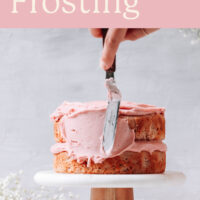 Vegan strawberry frosting being spread on vegan strawberry cake with fresh strawberries in the background