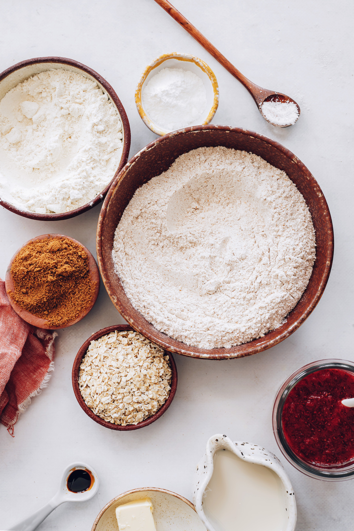 Bowls of spelt flour, whole wheat flour, rolled oats, coconut sugar, baking powder, and salt next to berry compote, dairy-free milk, vegan butter, and vanilla