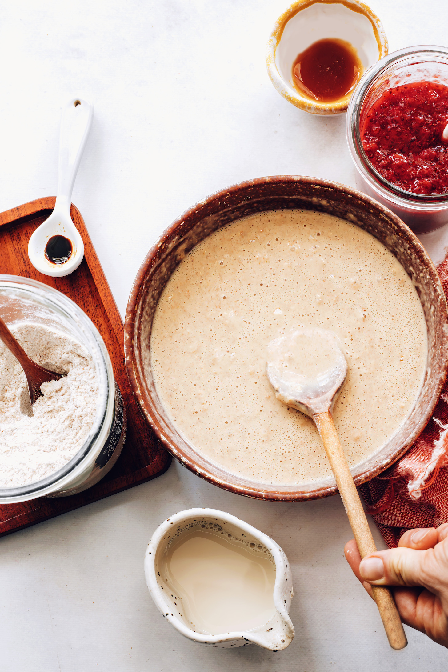Bowl of vegan pancake batter next to berry compote, pancake mix, almond milk, maple syrup, and vanilla