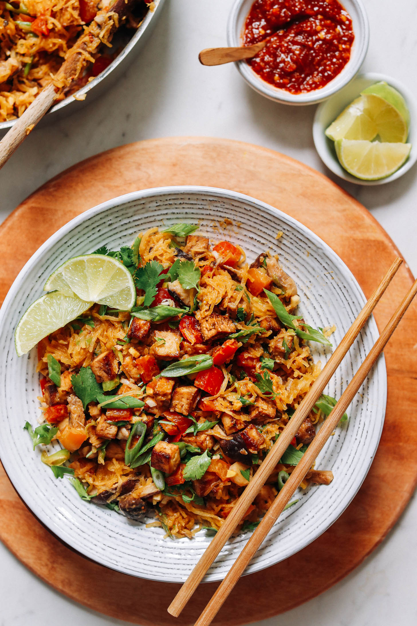 Chili garlic sauce and lime wedges beside a bowl of tofu and spaghetti squash stir-fry