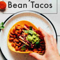 Plate and hand holding a vegan and gluten-free smoky bbq bean taco with guacamole on top