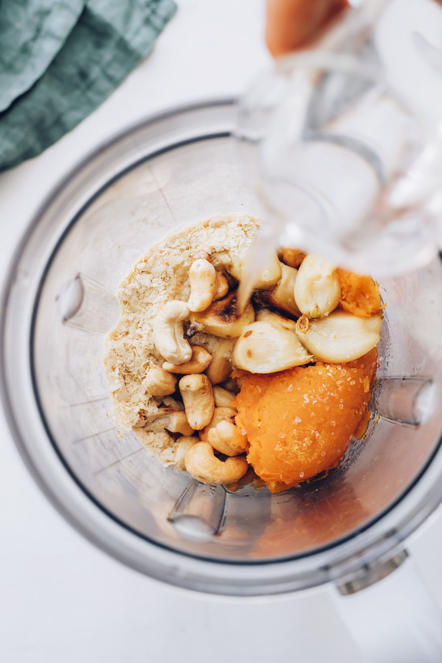 Pouring water into a blender with roasted squash, garlic, cashews, nutritional yeast, and salt