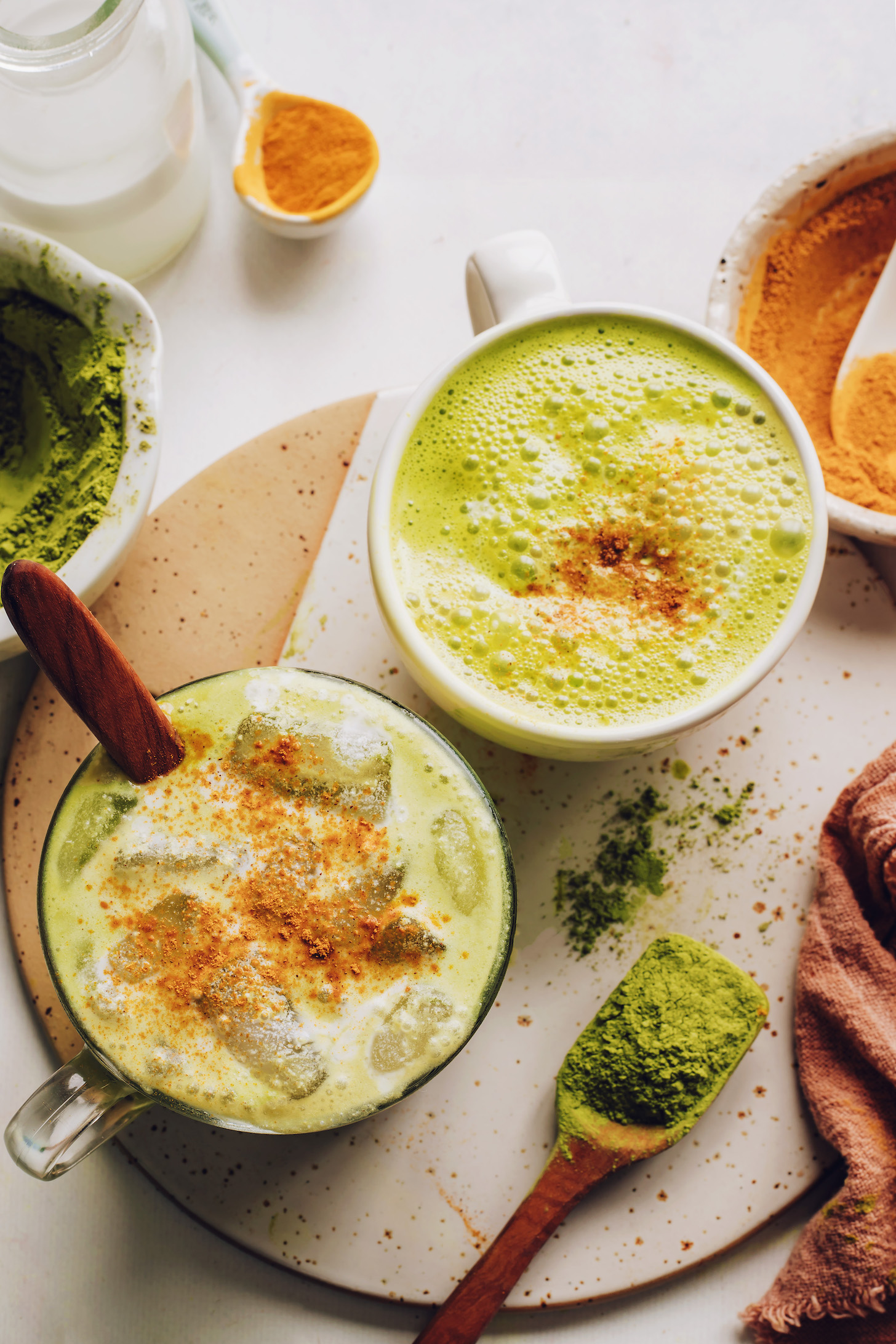 Mugs of iced and hot golden matcha lattes beside matcha powder and golden milk spices
