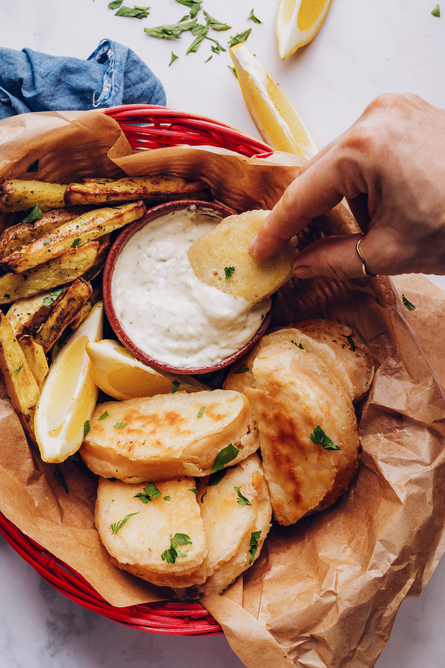 Basket of vegan fish and chips with a hand dipping one of the fish pieces in tartar sauce