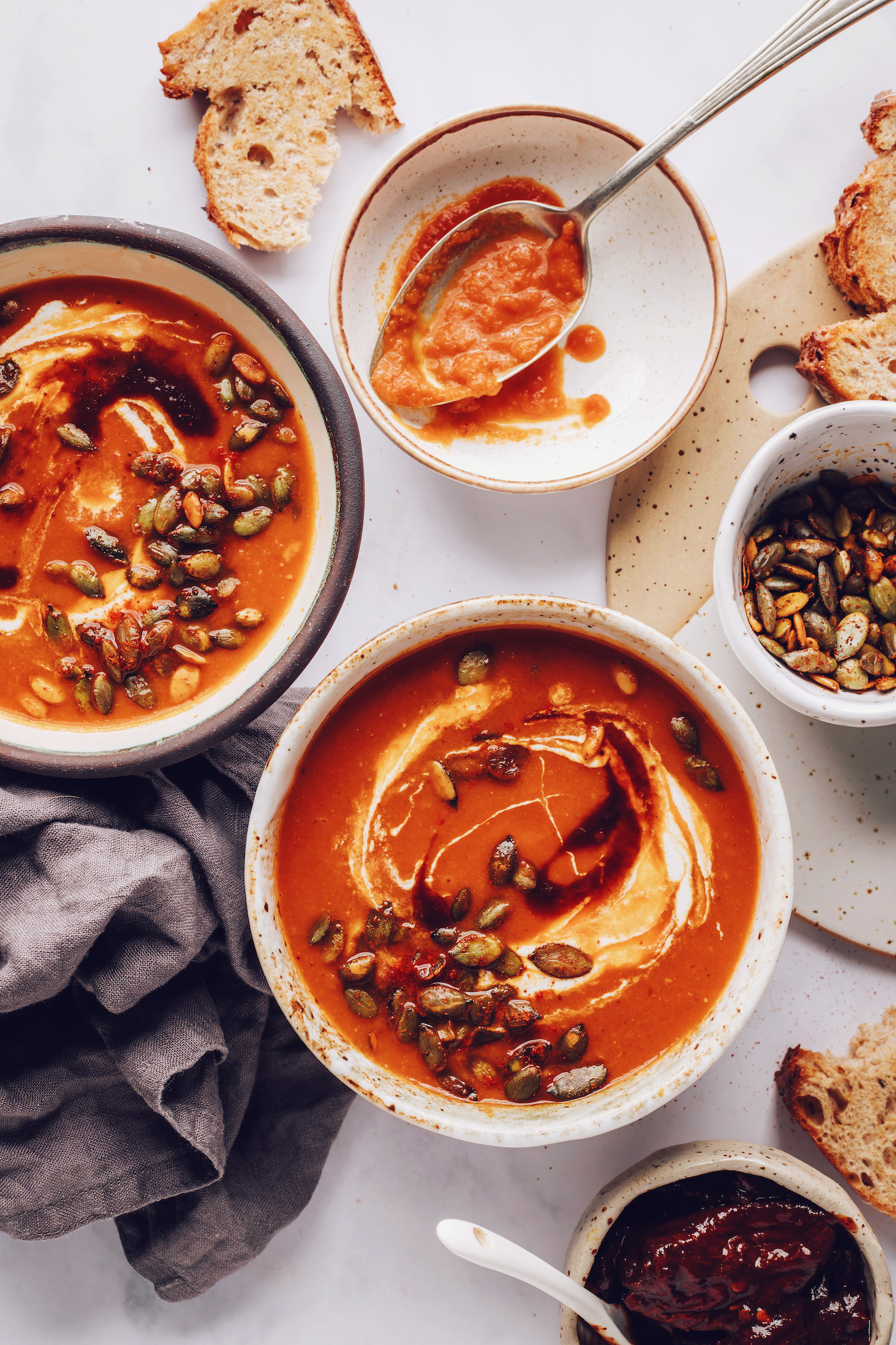 Slices of bread scattered around bowls of spicy tomato soup with coconut milk and pepitas
