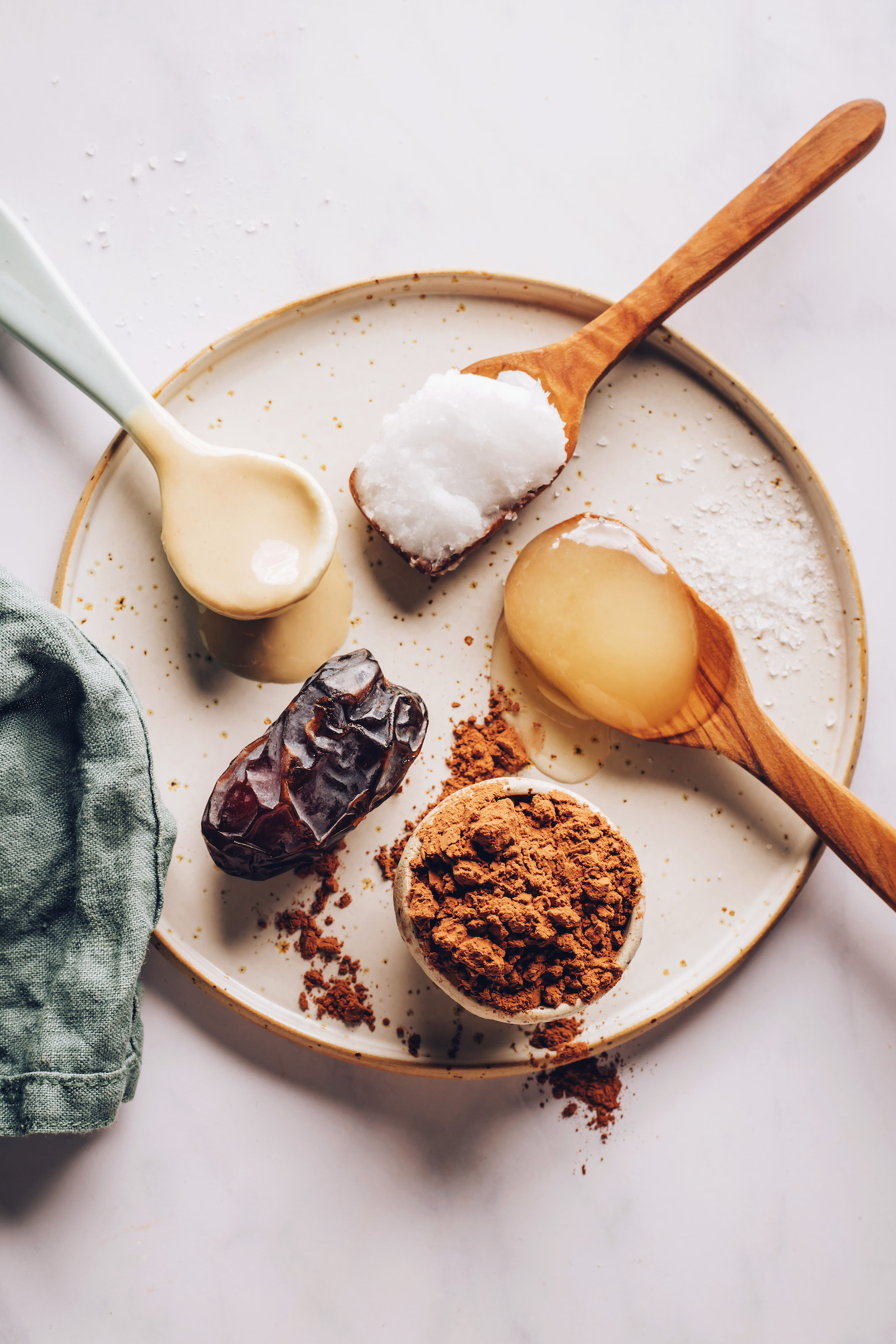Spoonfuls of tahini, coconut oil, and honey next to a medjool date and bowl of cocoa powder