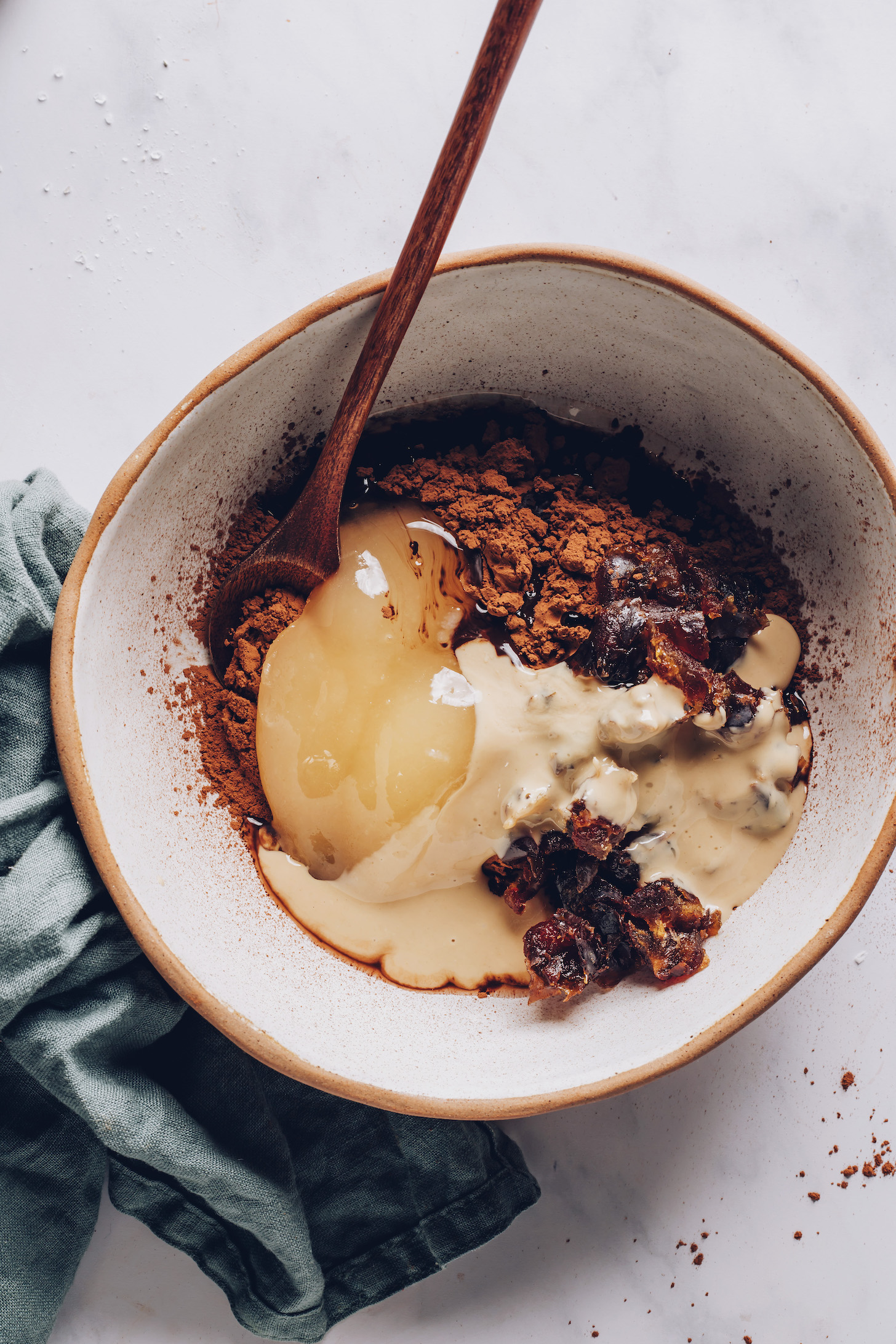 Wooden spoon in a bowl of cocoa powder, cacao powder, tahini, honey, melted coconut oil, and dates
