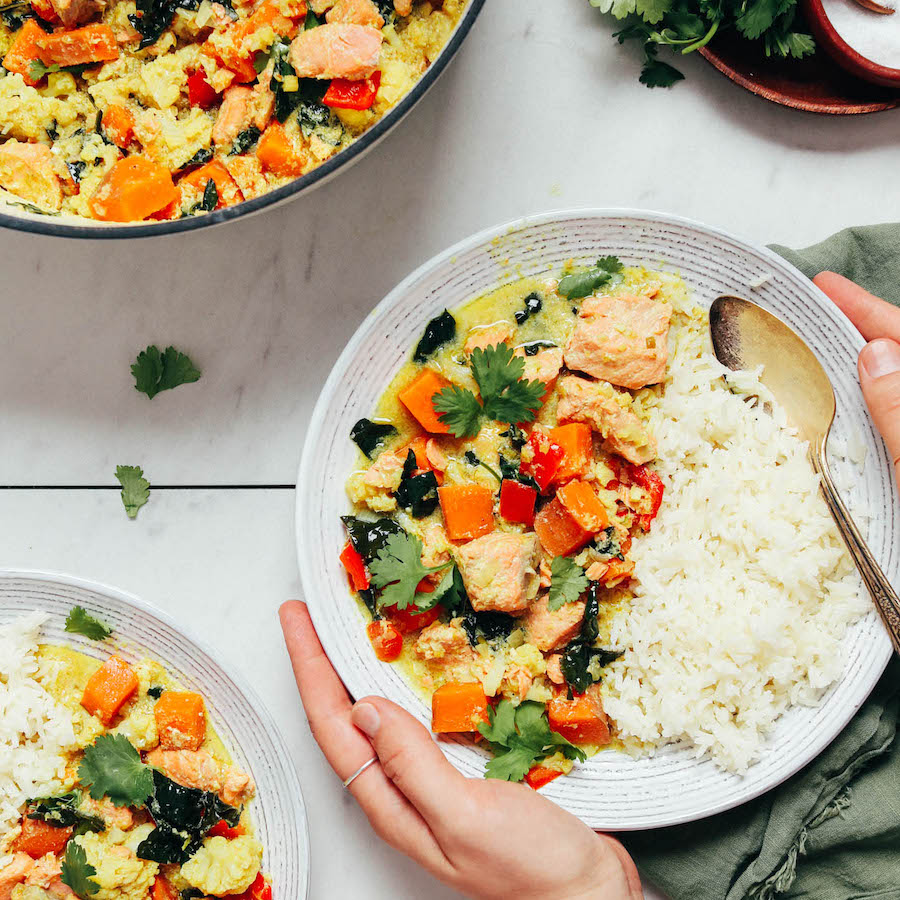 Hands holding a bowl of salmon green curry with white rice
