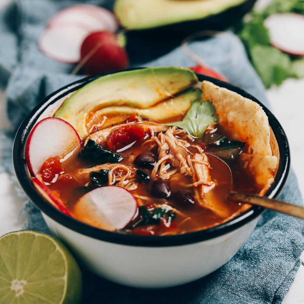 Bowl of chicken tortilla soup topped with avocado, sliced radish, and tortilla chips
