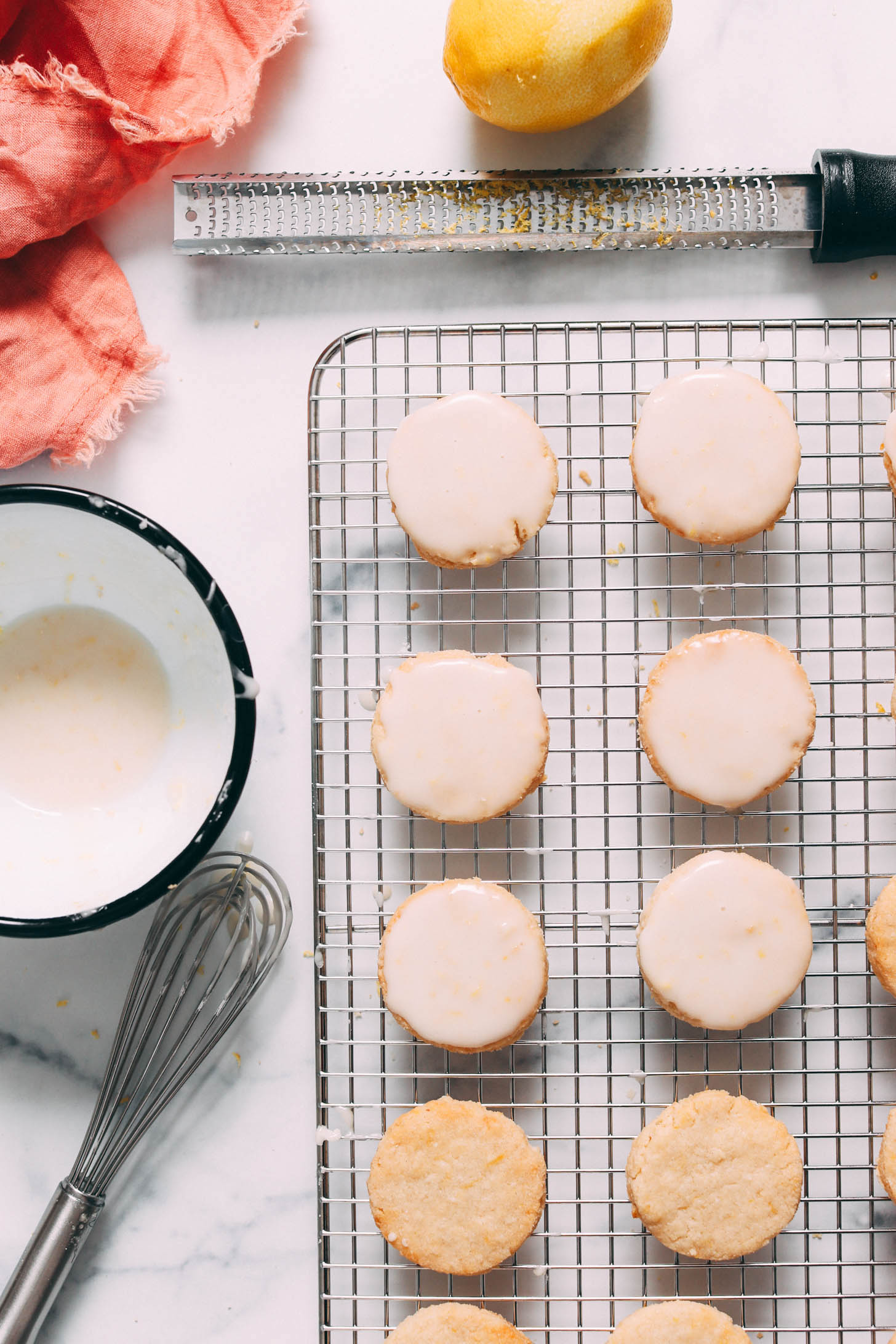 Glazed lemon cookies on a cooling rack next to a bowl of glaze and a zested lemon