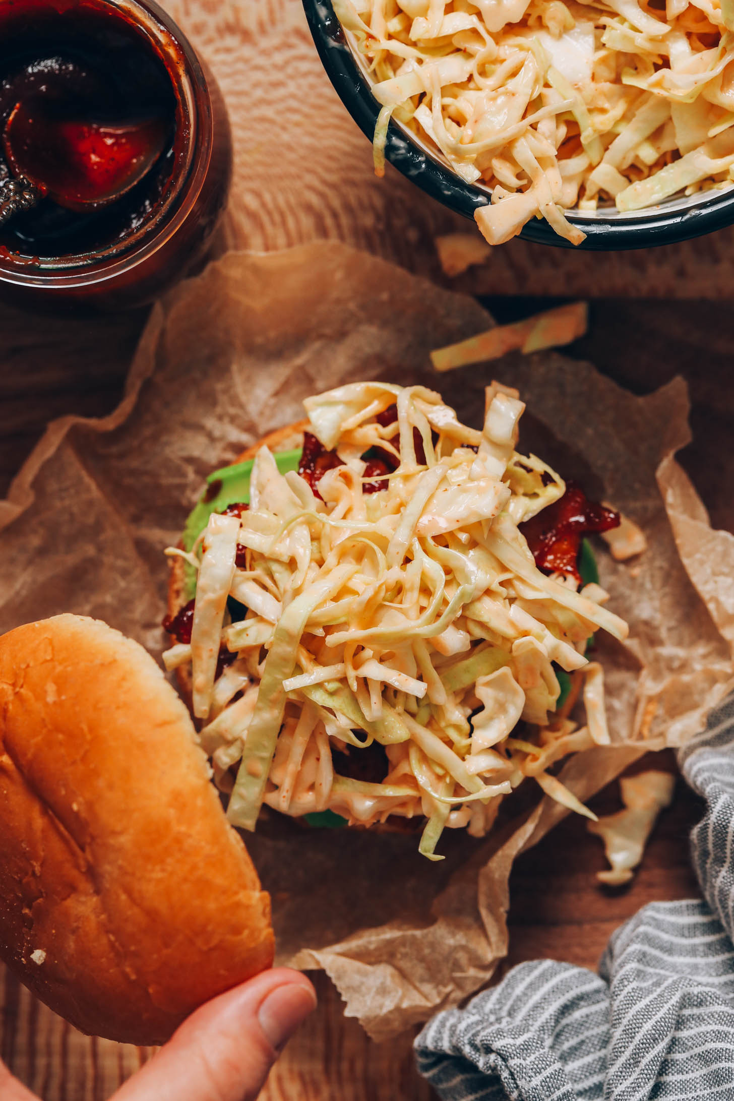 BBQ sauce and slaw next to a hand holding a bun over a sandwich
