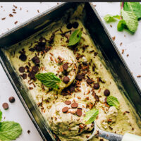 Ice cream scooper in a container of vegan no-churn mint chocolate chip ice cream with mint leaves and chocolate chips on top