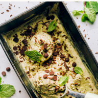 Ice cream scooper in a container of vegan no-churn mint chocolate chip ice cream with mint leaves and chocolate chips on top