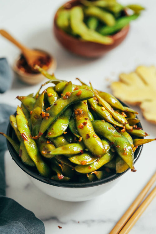Bowl of spicy garlic edamame pods next to ginger and chili flakes