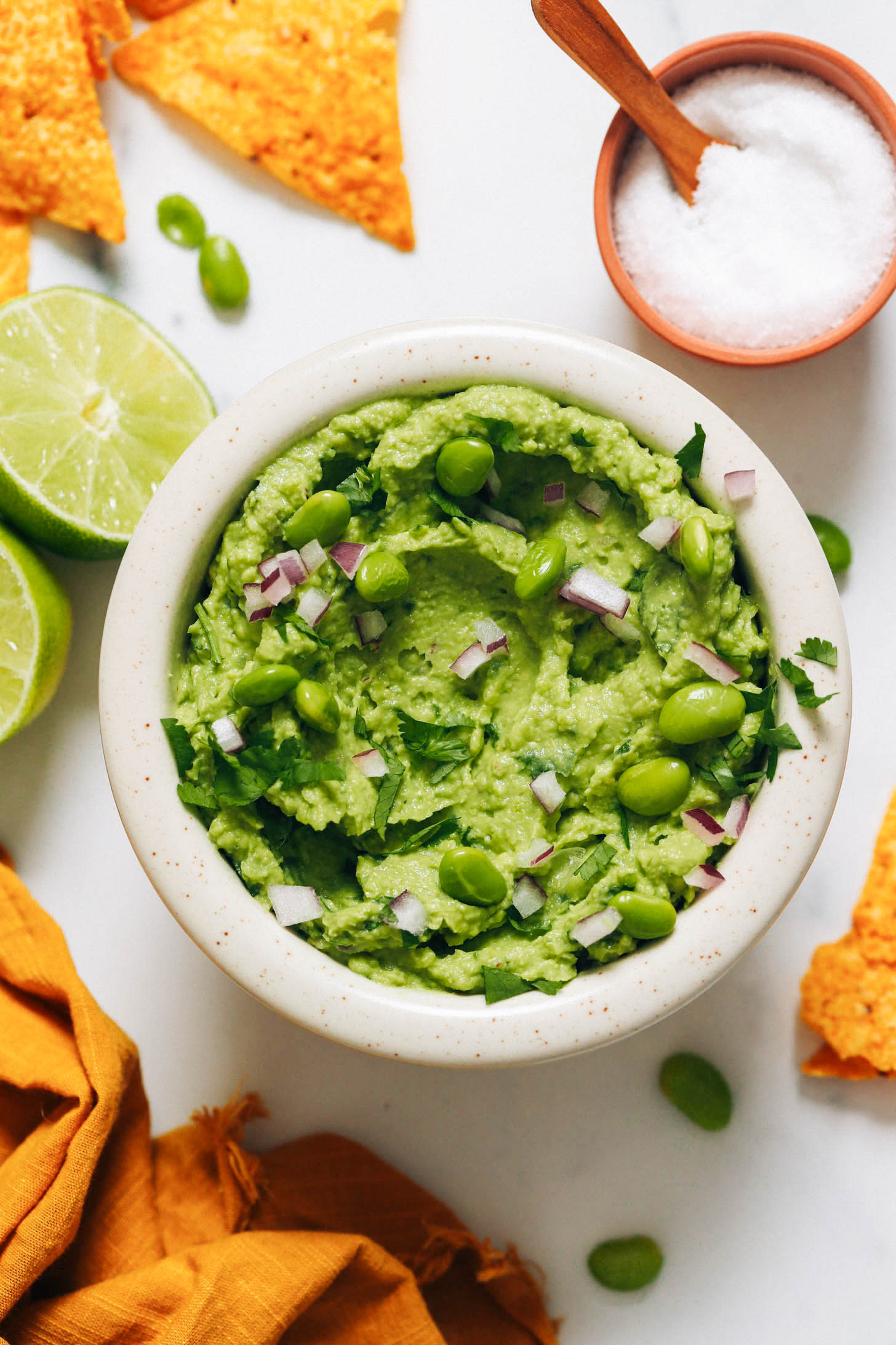 Salt, tortilla chips, edamame, and limes surrounding a bowl of edamame guacamole