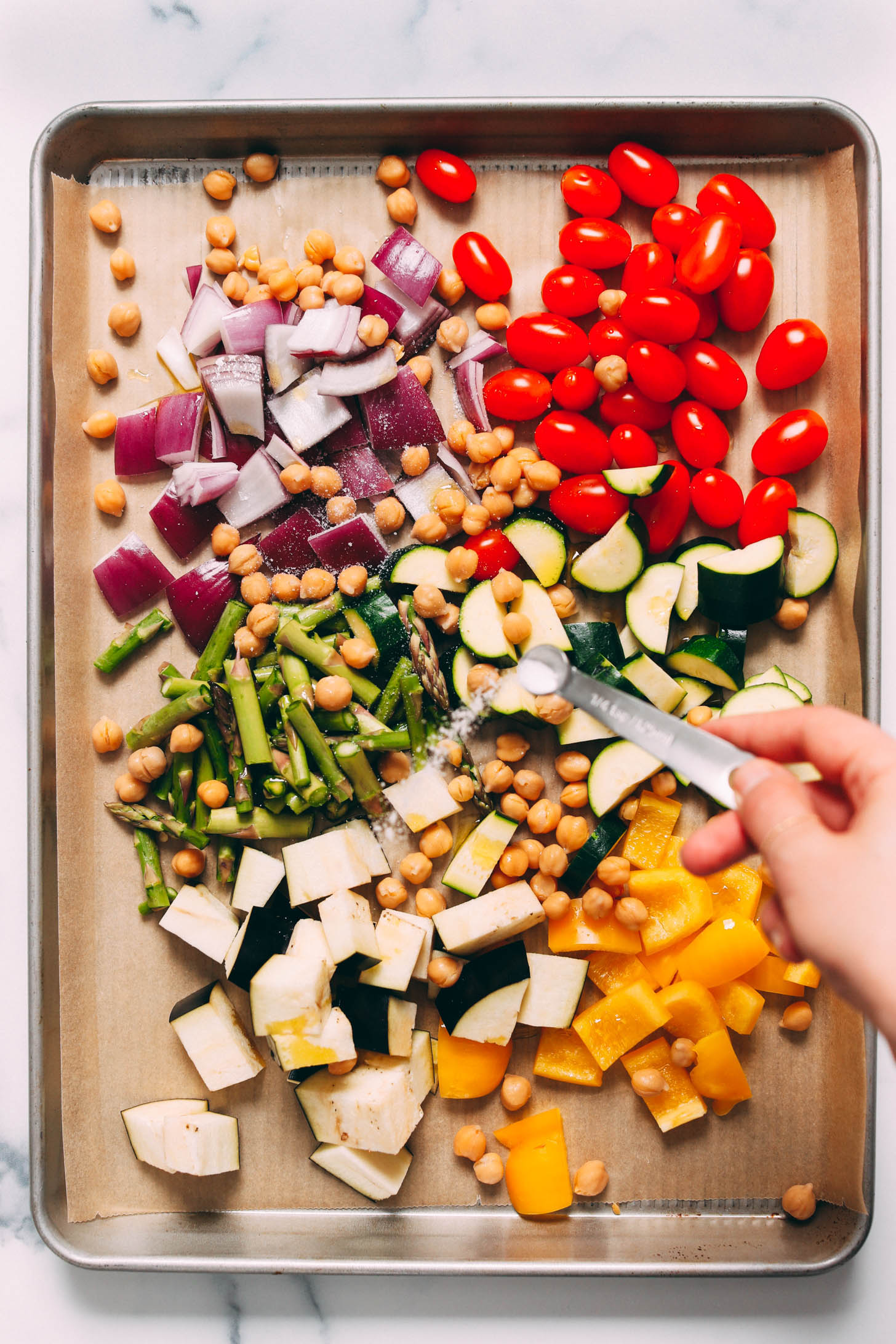 Sprinkling salt onto a baking sheet of chopped red onion, chickpeas, cherry tomatoes, asparagus, zucchini, eggplant, and bell pepper