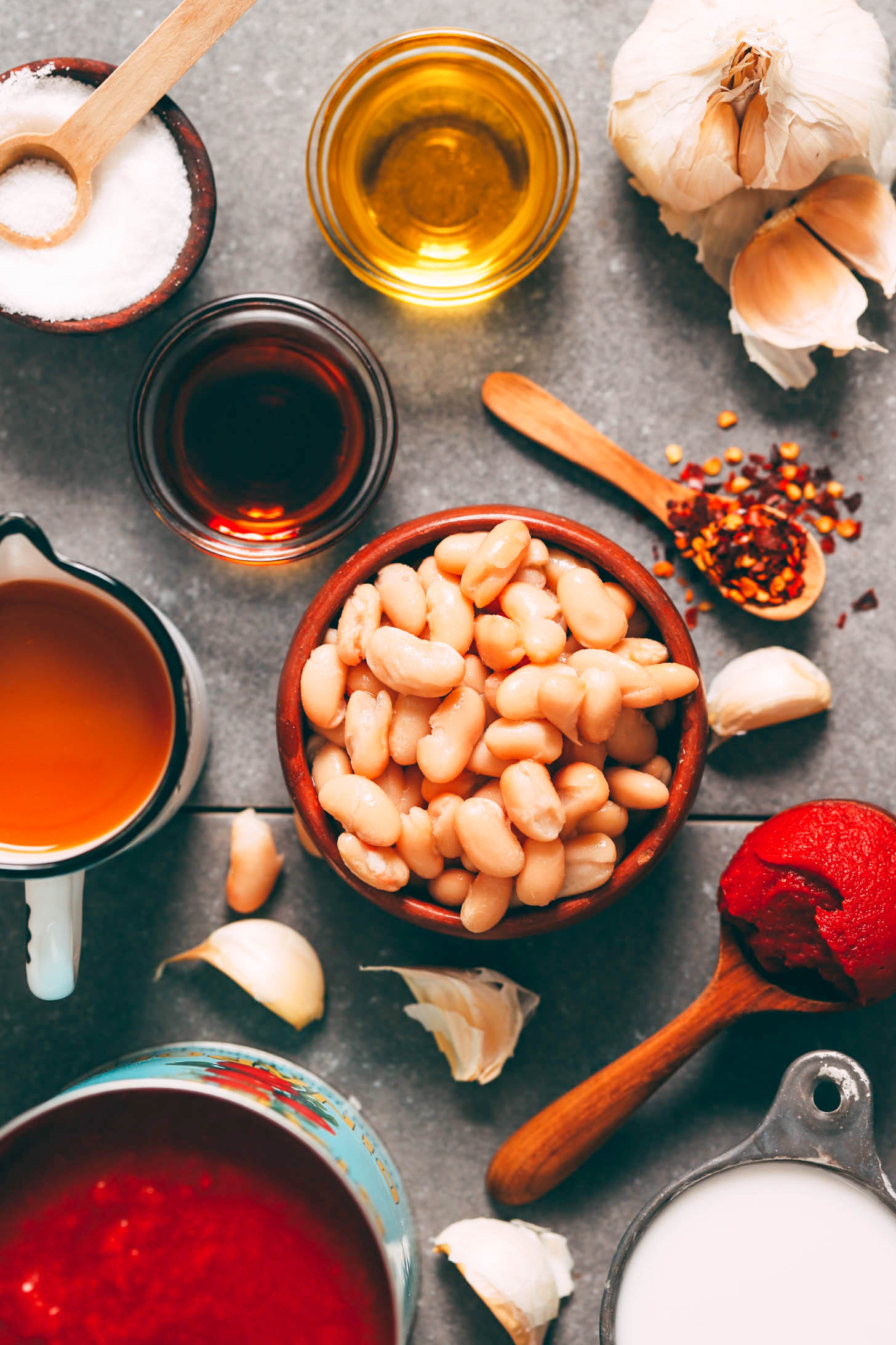 Salt, olive oil, garlic, red pepper flakes, tomato paste, coconut milk, crushed tomatoes, vegetable broth, and maple syrup around bowl of beans