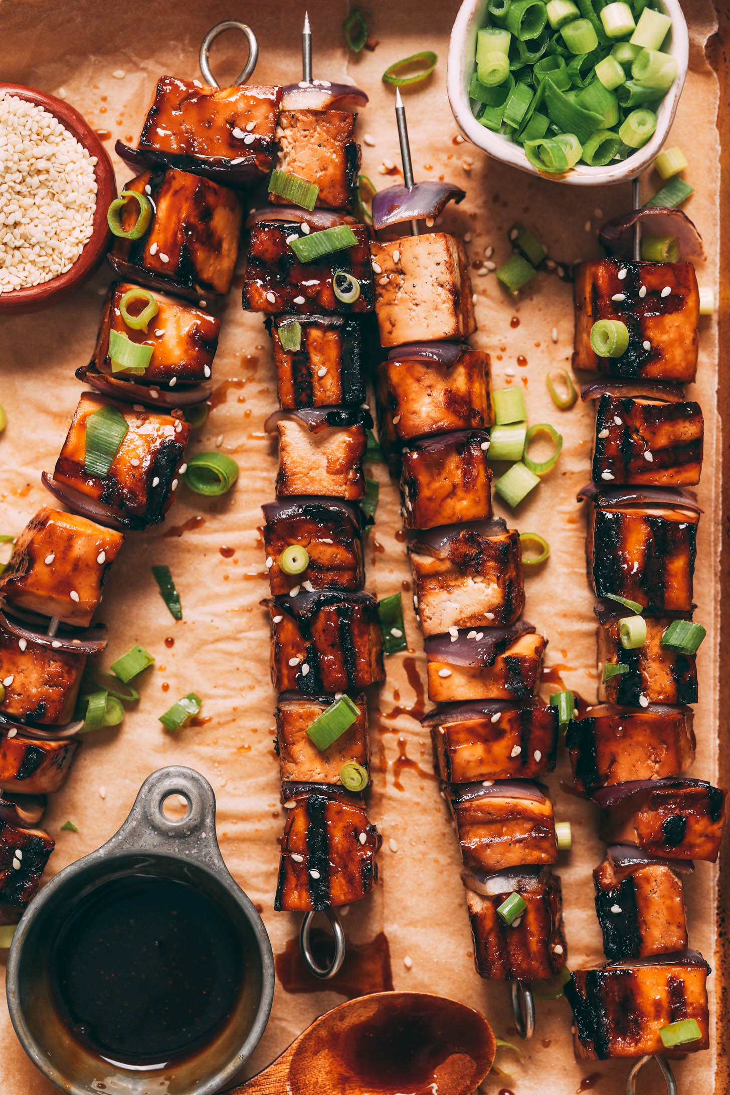 Teriyaki sauce, sesame seeds, and green onions on a baking sheet next to teriyaki tofu skewers