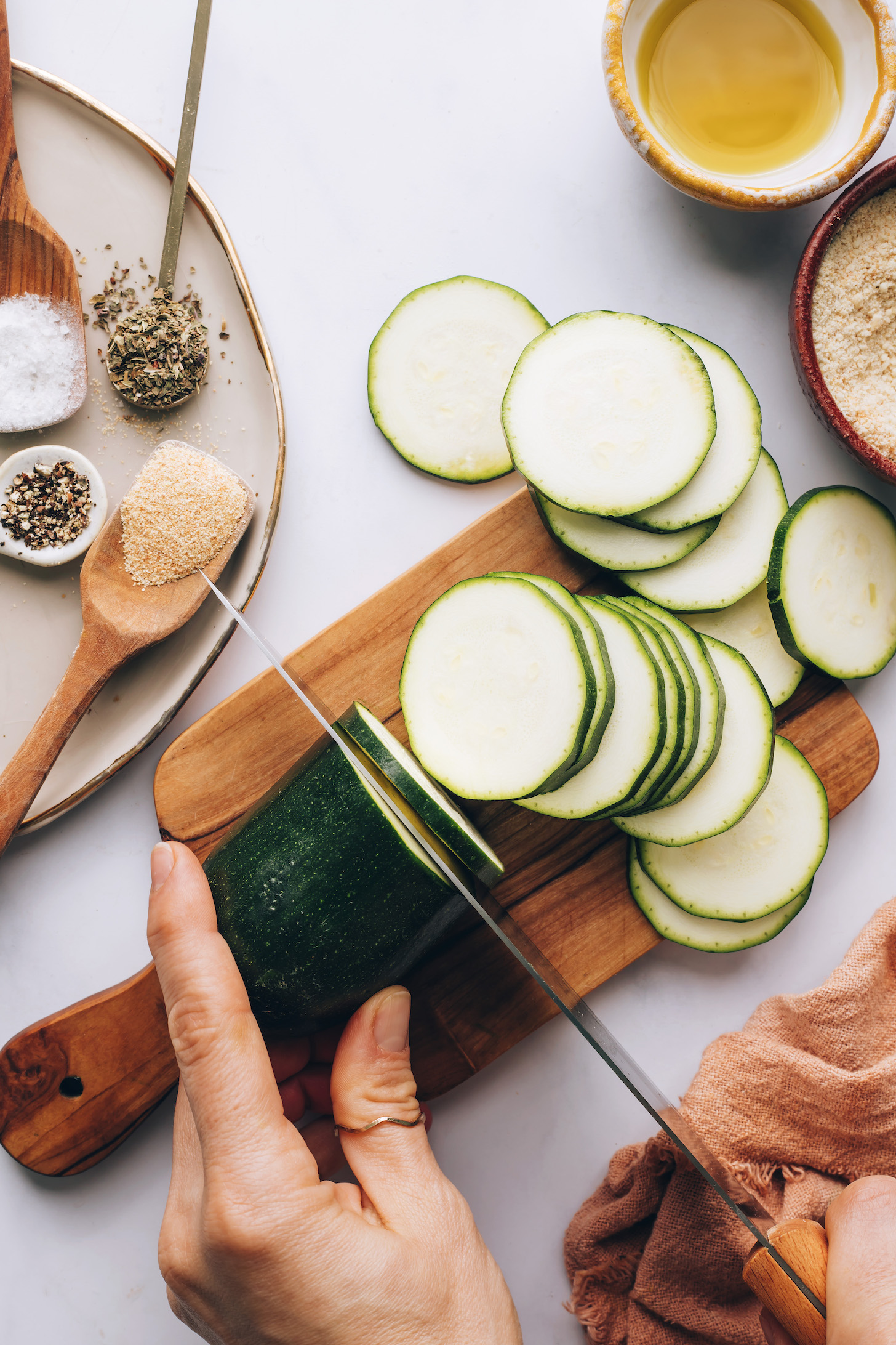 Thinly slicing a zucchini with oil, vegan parmesan, and spices next to it