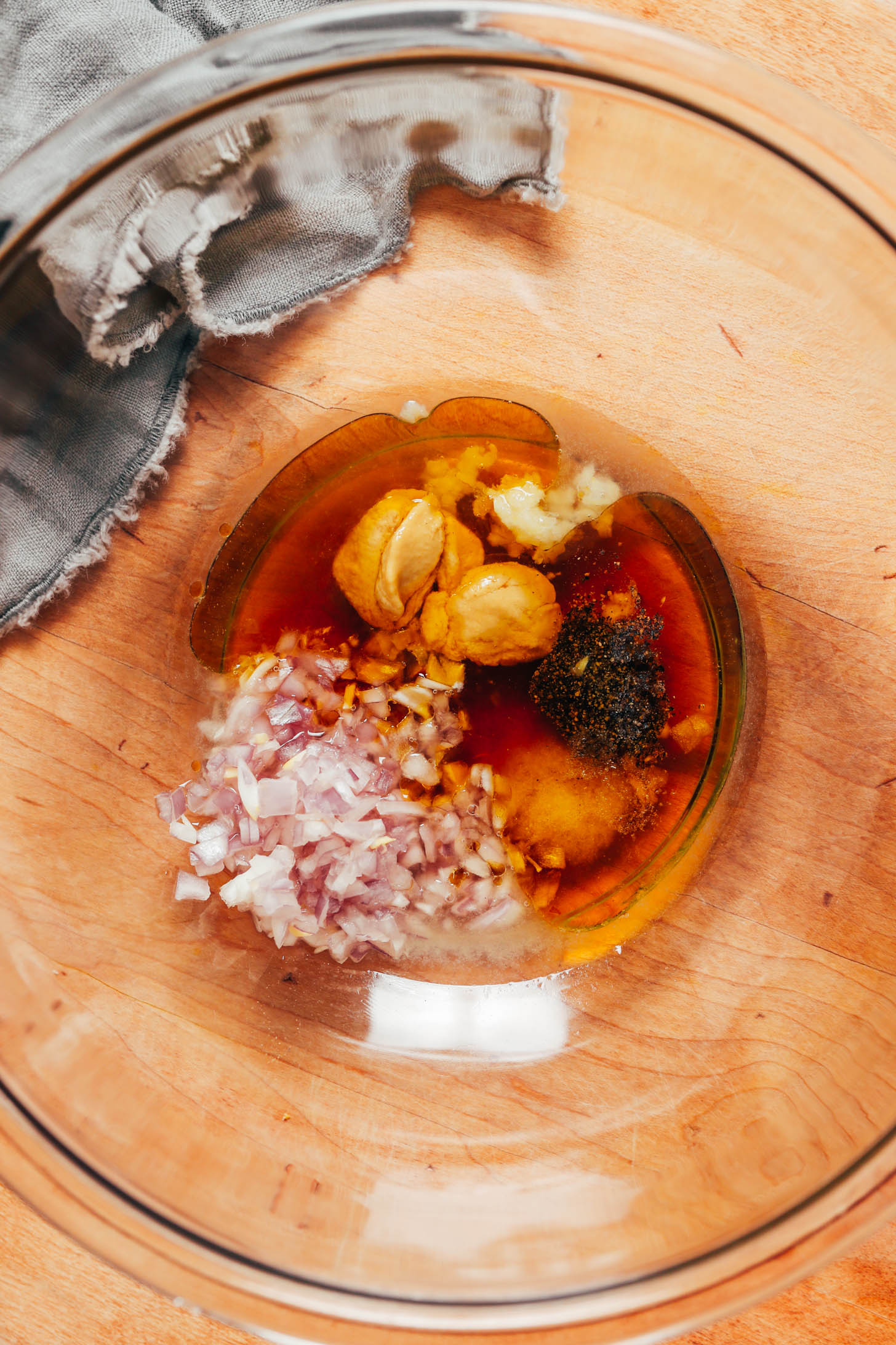 Glass mixing bowl with olive oil, lemon juice, garlic, Dijon mustard, maple syrup, shallot, salt, and pepper